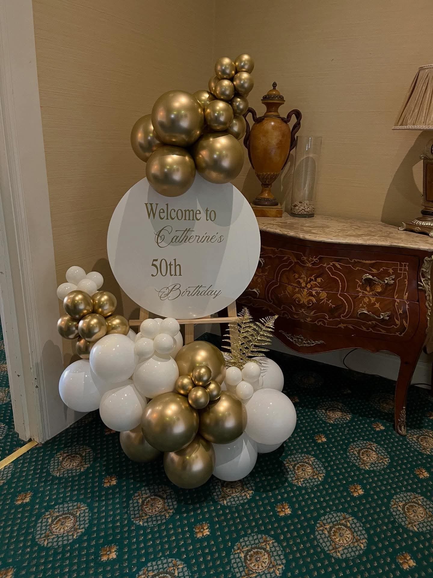 Balloon arrangement with gold, white, and metallic balloons surrounding a sign that reads 'Welcome to Catherine's 50th Birthday,' set in an indoor space with an antique wooden dresser and decorative items.