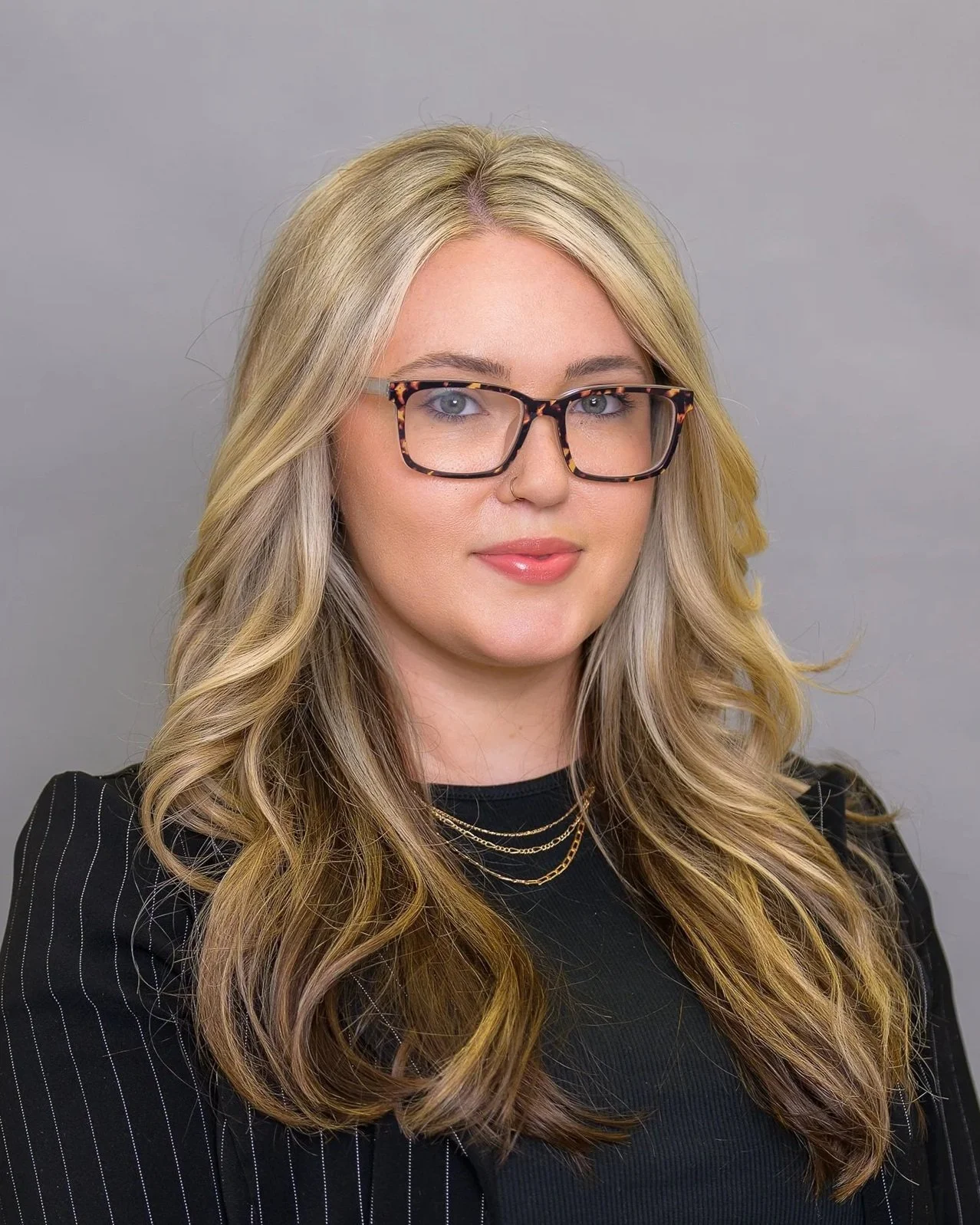 A woman with wavy blonde hair and glasses, smiling at the camera, wearing a black top and a gold necklace, against a plain grey background.