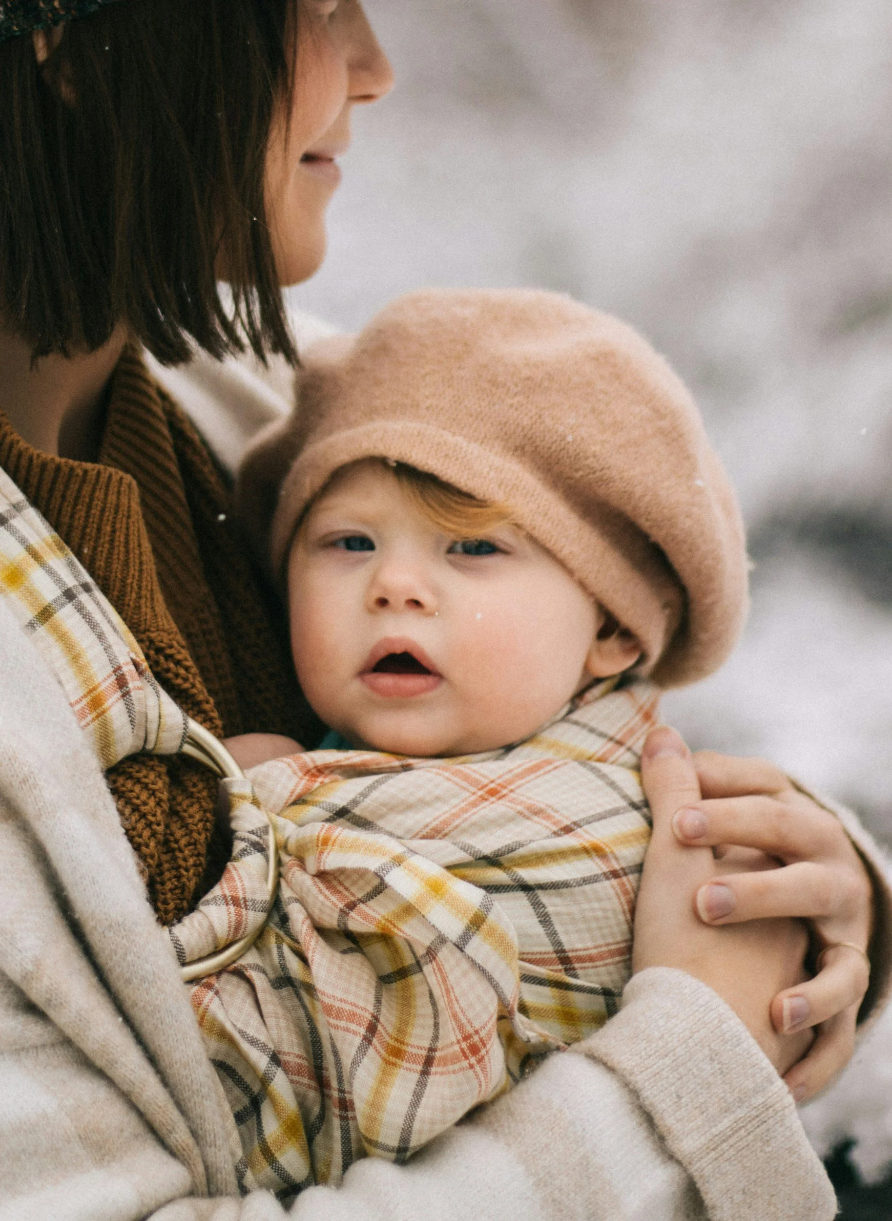 A woman holding a young child with a beige beret, wrapped in a checked blanket, in a winter outdoor setting.