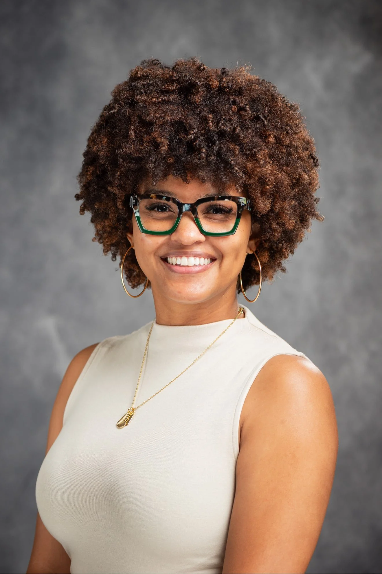 A woman with curly hair, wearing glasses, hoop earrings, a gold necklace, and a sleeveless denim top, smiling against a light purple background.