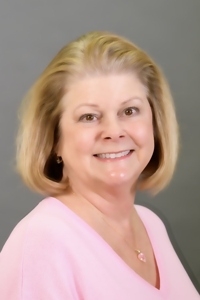 A woman with shoulder-length blonde hair, wearing a pink top and jewelry, smiling against a gray background.