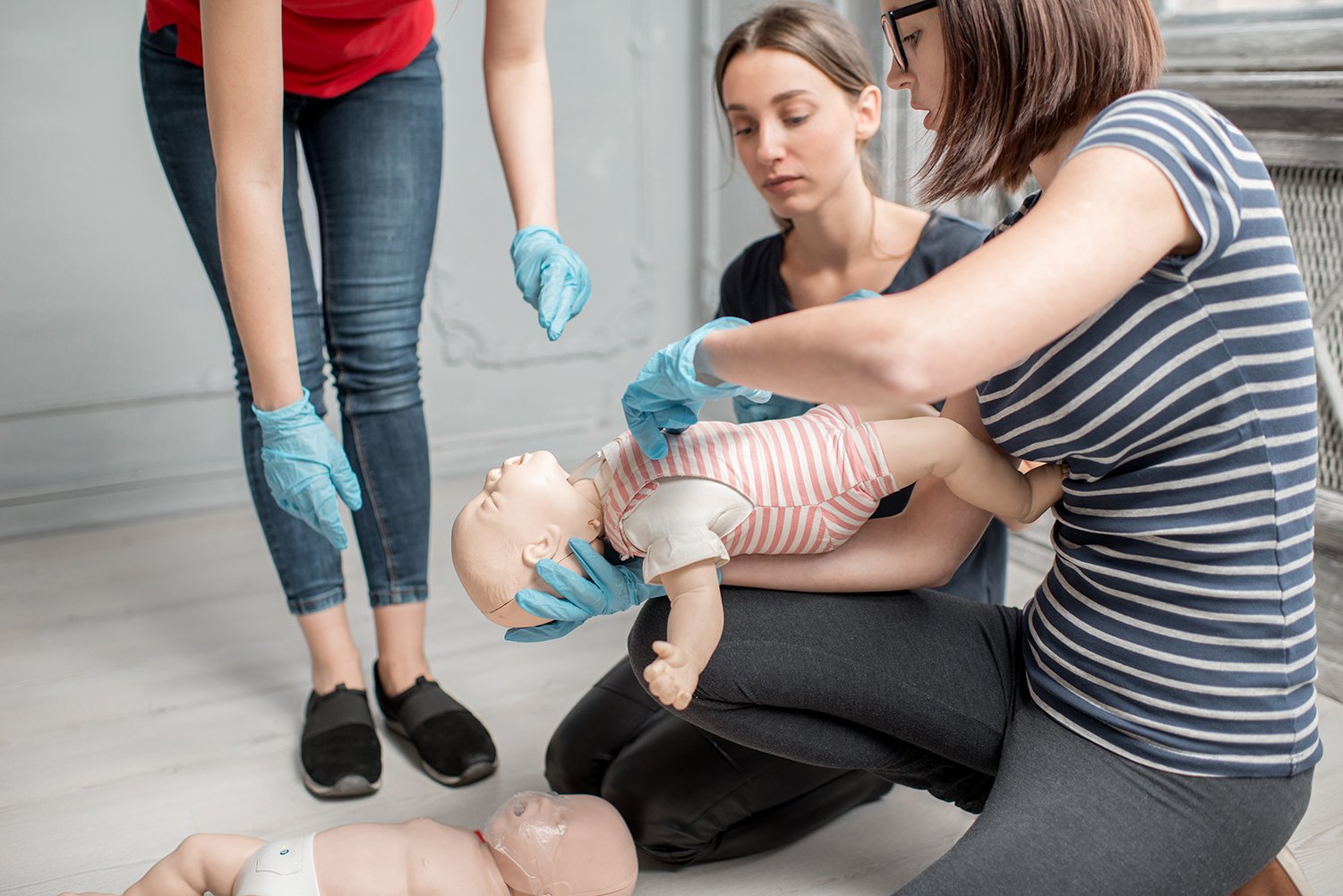 Three women practicing infant CPR on a mannequin baby, using gloves, indoors.