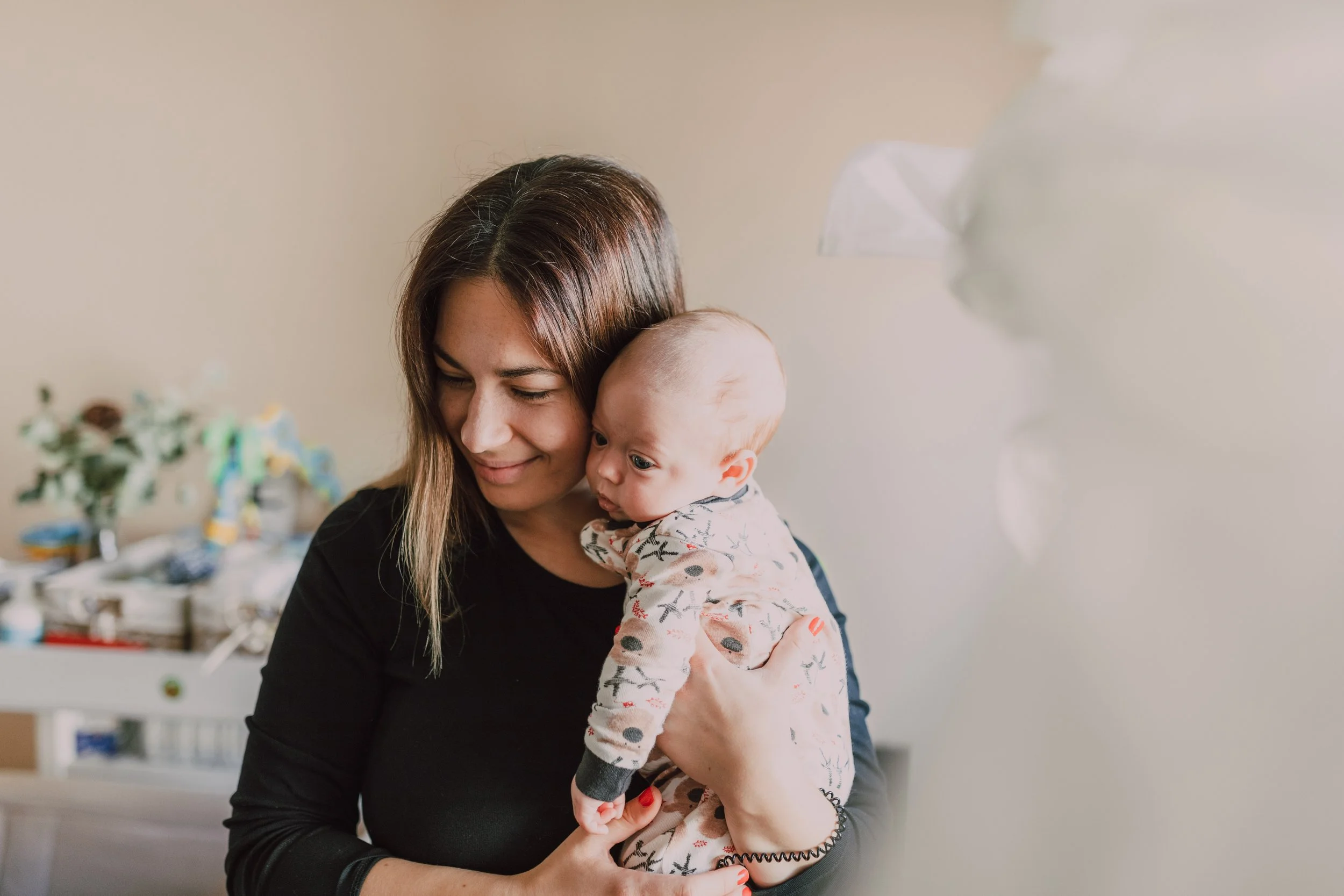 A woman holding a baby, both smiling and looking down, in a warmly lit room with blurred background.