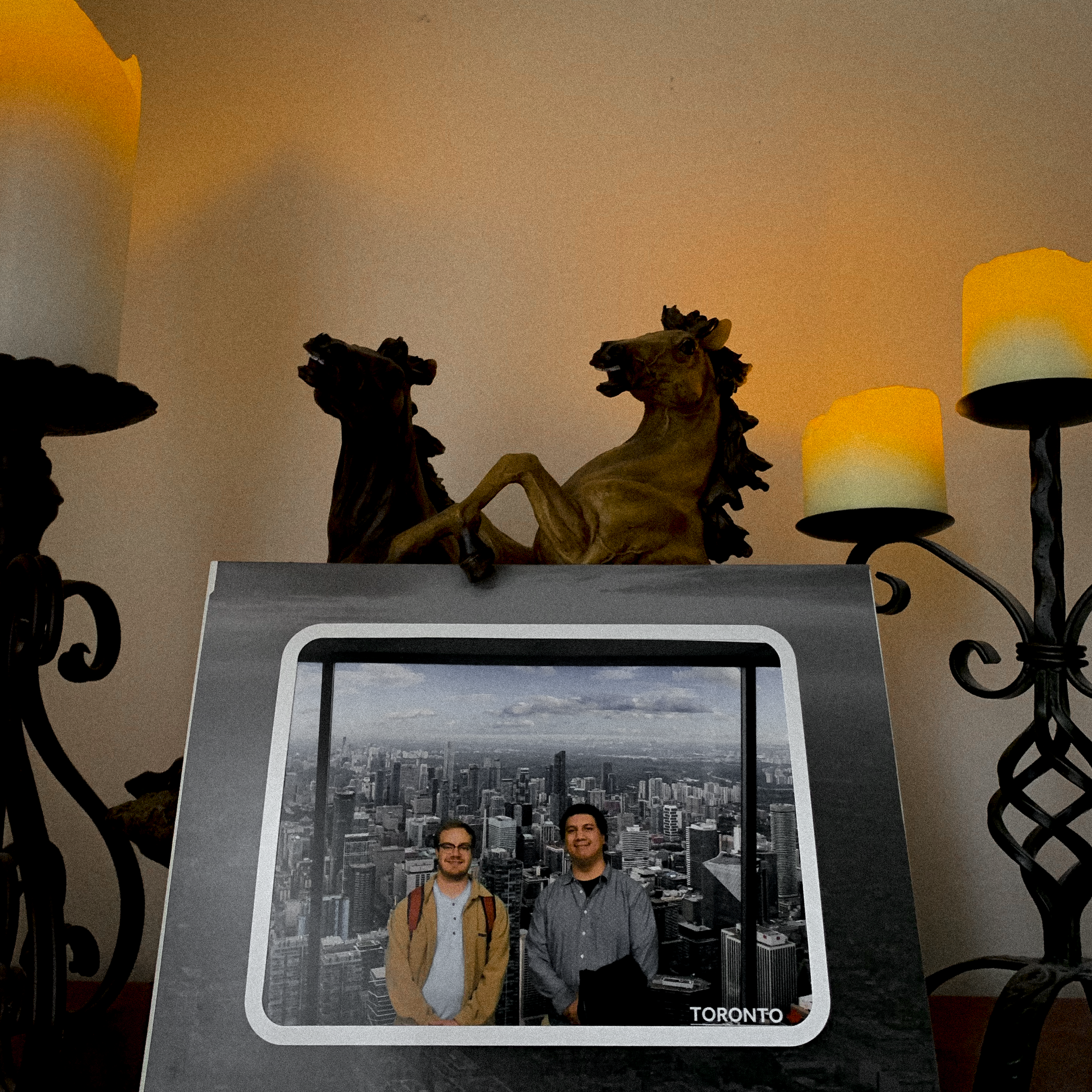Une table avec deux statues de chevaux en bois, deux bougies sur des candélabres en métal, une carte postale de FLATTOP et Simon Desro avec la ville de Toronto en arrière-plan.