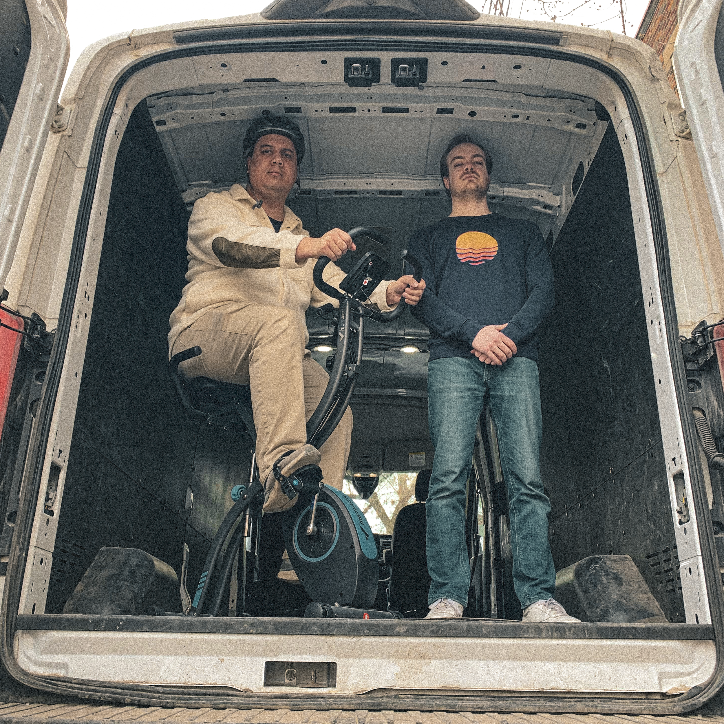 FLATTOP et Simon Desro dans une camionnette, l'un debout à côté d'un vélo d'exercice, l'autre assis dessus, regardant vers l'objectif.