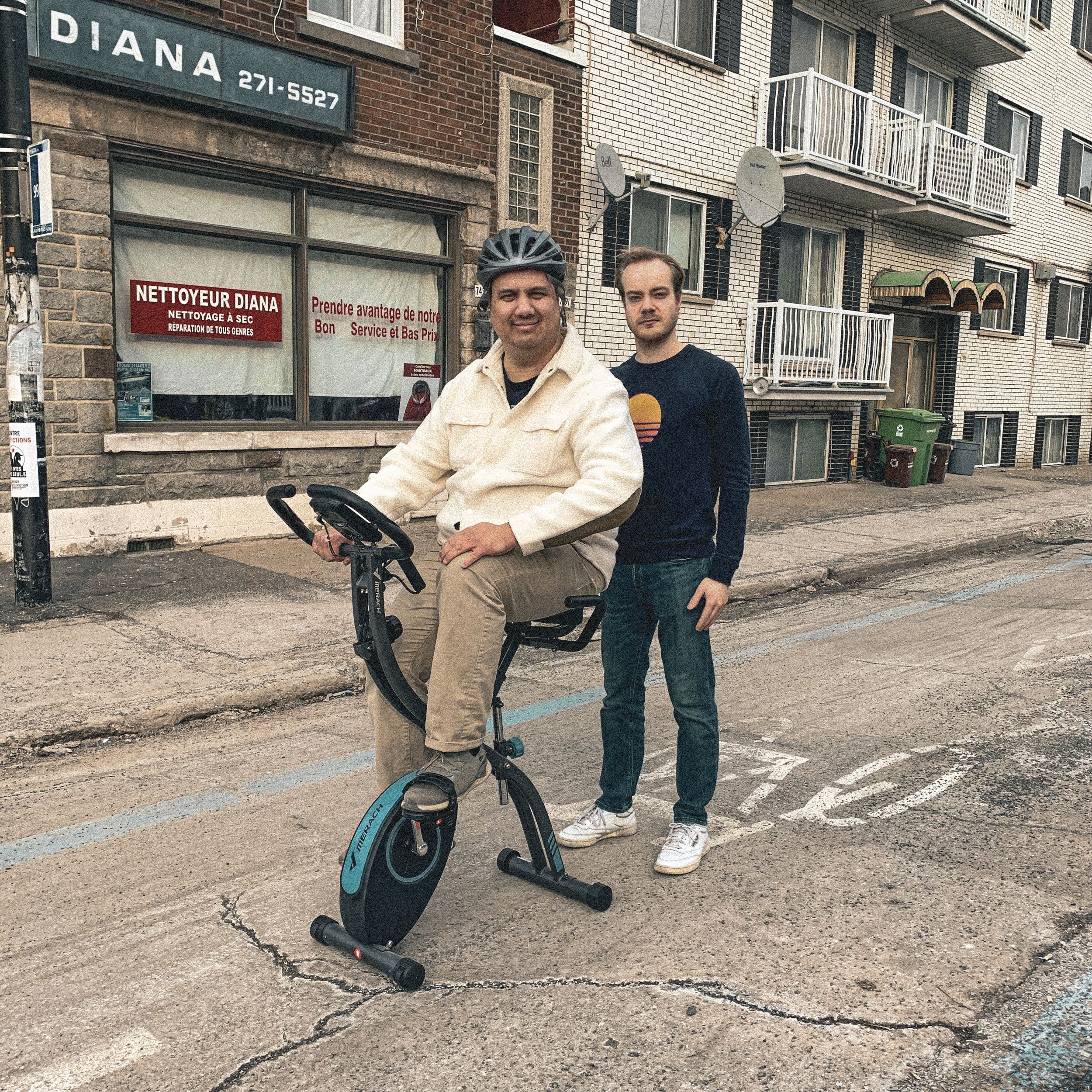 FLATTOP et Simon Desro debout sur une rue, l'un assis sur un vélo d'exercice et portant un casque. En arrière-plan, un immeuble résidentiel et un commerce avec une enseigne en français.
