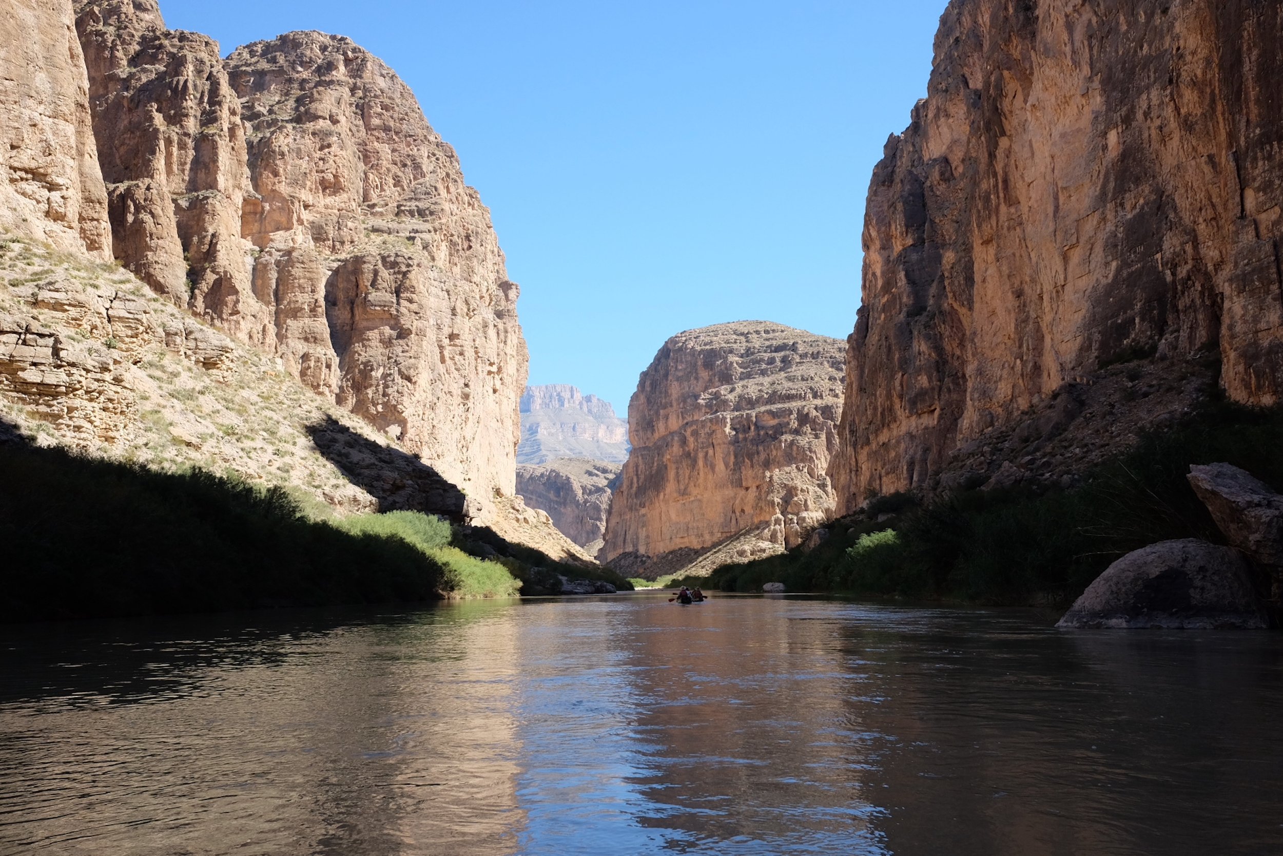 A narrow river flowing through a deep canyon with tall, rugged rock formations on both sides under a clear blue sky, with a small boat floating on the water.