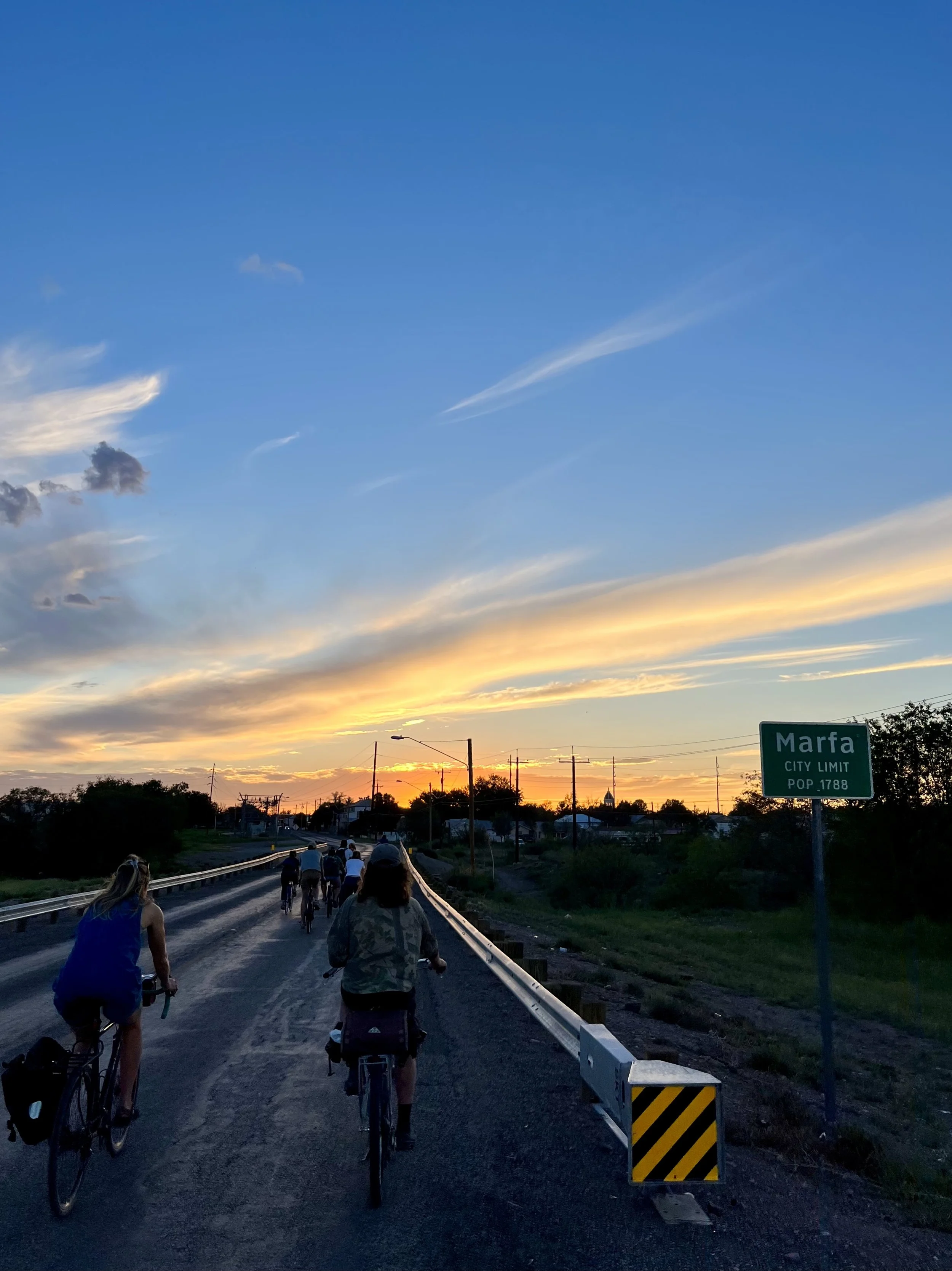 Bicyclists riding on a rural road at sunset near Marfa, Texas, with a colorful sky and a city limit sign in the background.