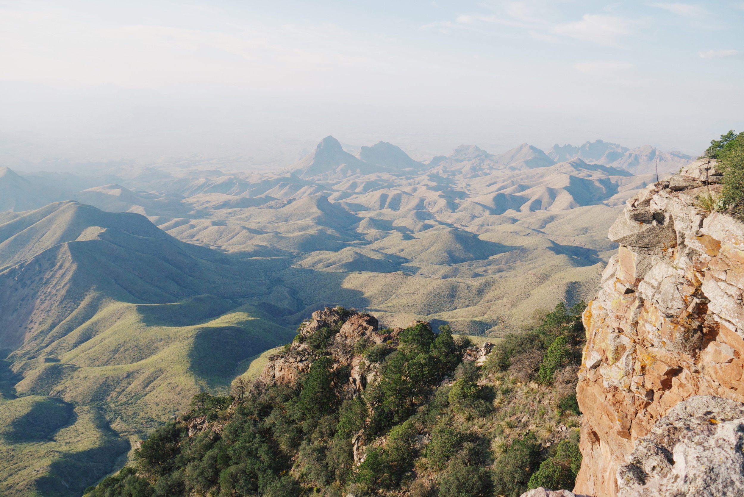 Scenic view of rolling green hills and mountains with a rocky cliff in the foreground under a partly cloudy sky.