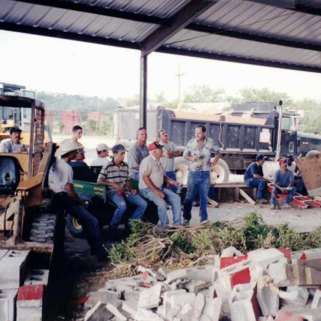 Group of people sitting and standing inside a construction or industrial site, with construction equipment and trucks in the background under a roofed structure.