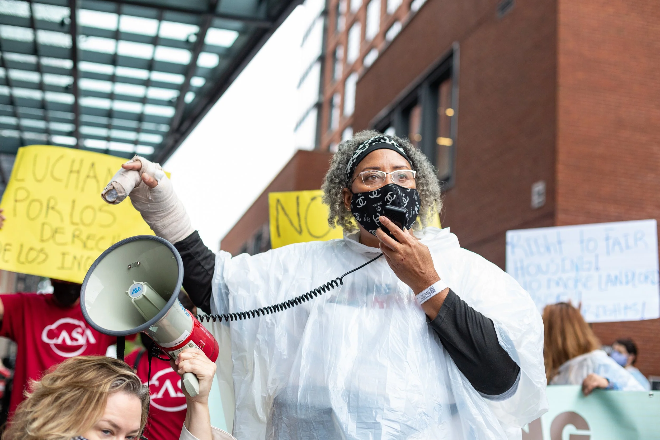 A brown skinned person with curly light brown hair, wearing glasses and a clear rain poncho, speaking into a bullhorn microphone. There are protestors with signs in the background.