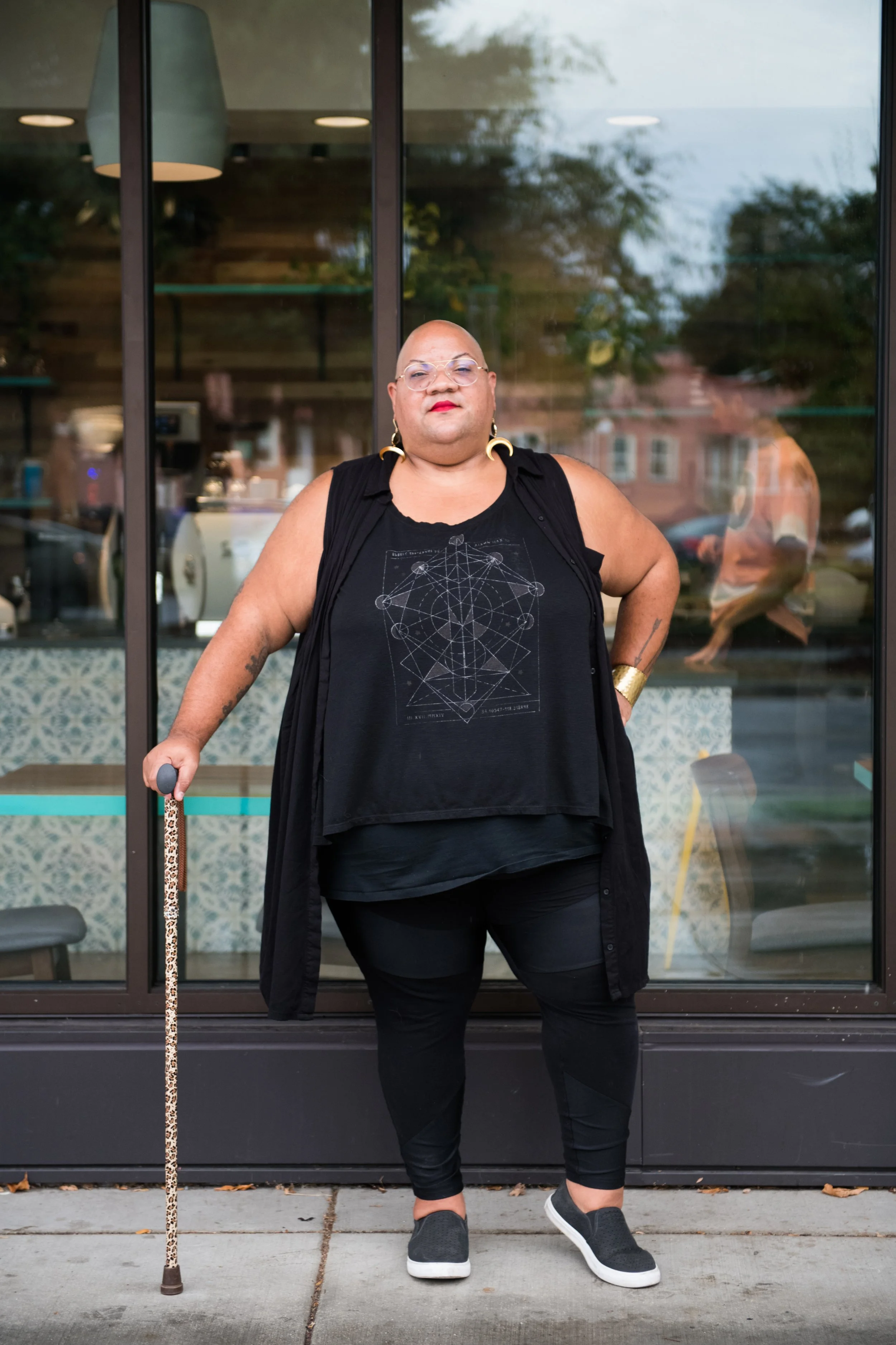 A bald, brown-skinned person wearing glasses and a black dress, standing with a cane.