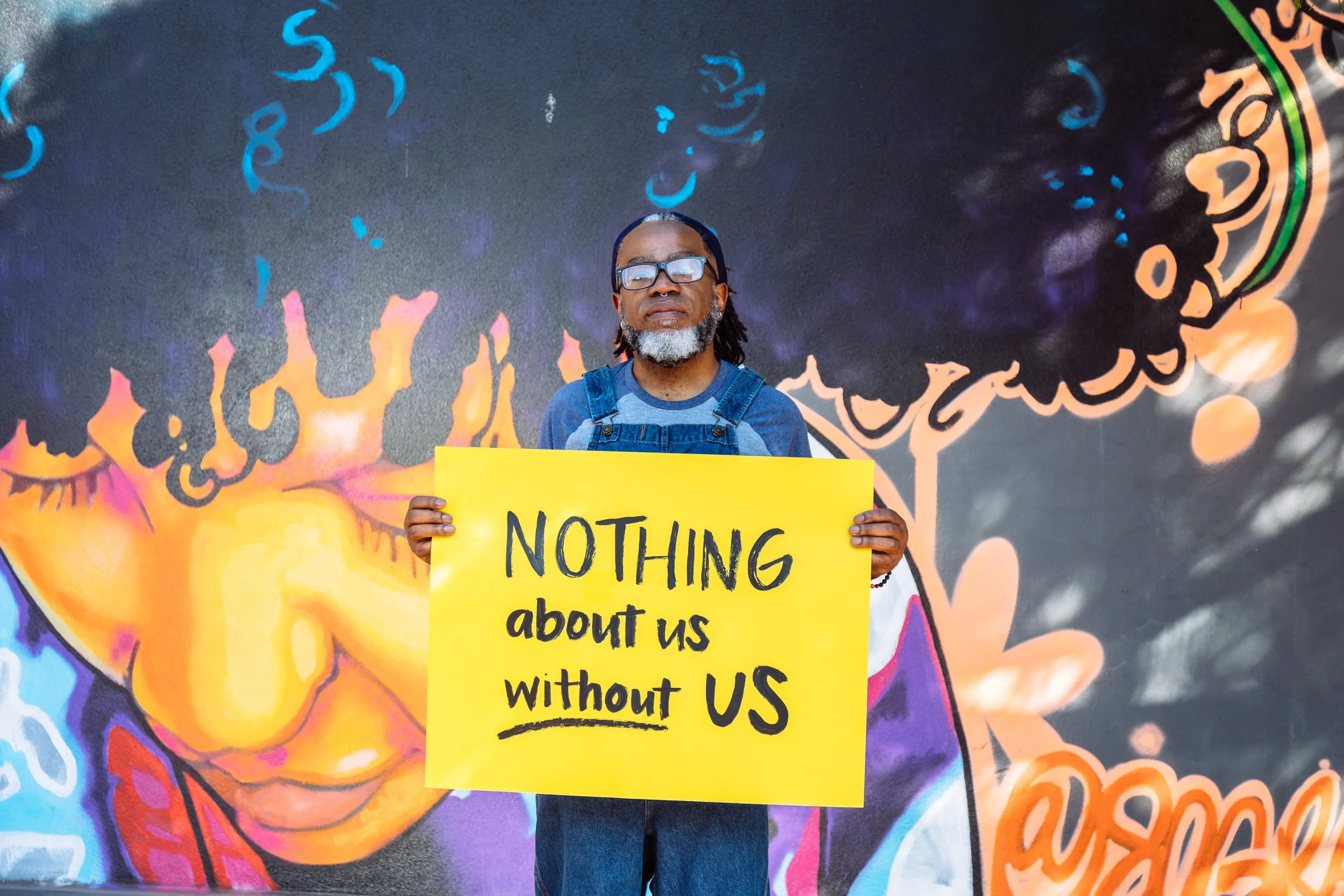 A dark-skinned person with a gray beard, gray locs, and sunglasses, holds a yellow sign that read “Nothing About Us Without Us”. They stand before a colorful mural of a person with curly hair.