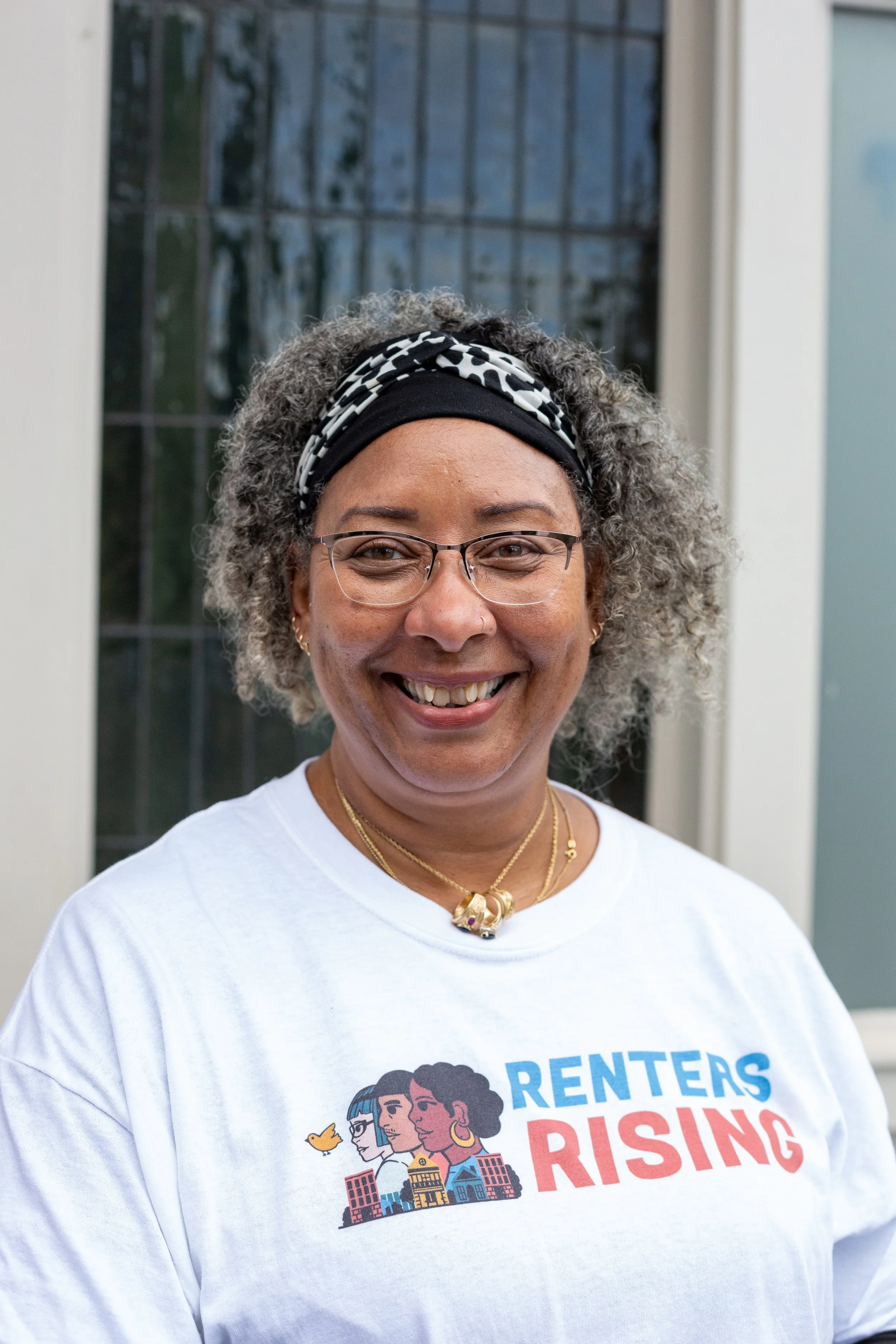 A brown-skinned person with chin-length silver hair and glasses, smiling.  They are wearing a white t-shirt with the words “Renters Rising” with an illustration of multiracial people.
