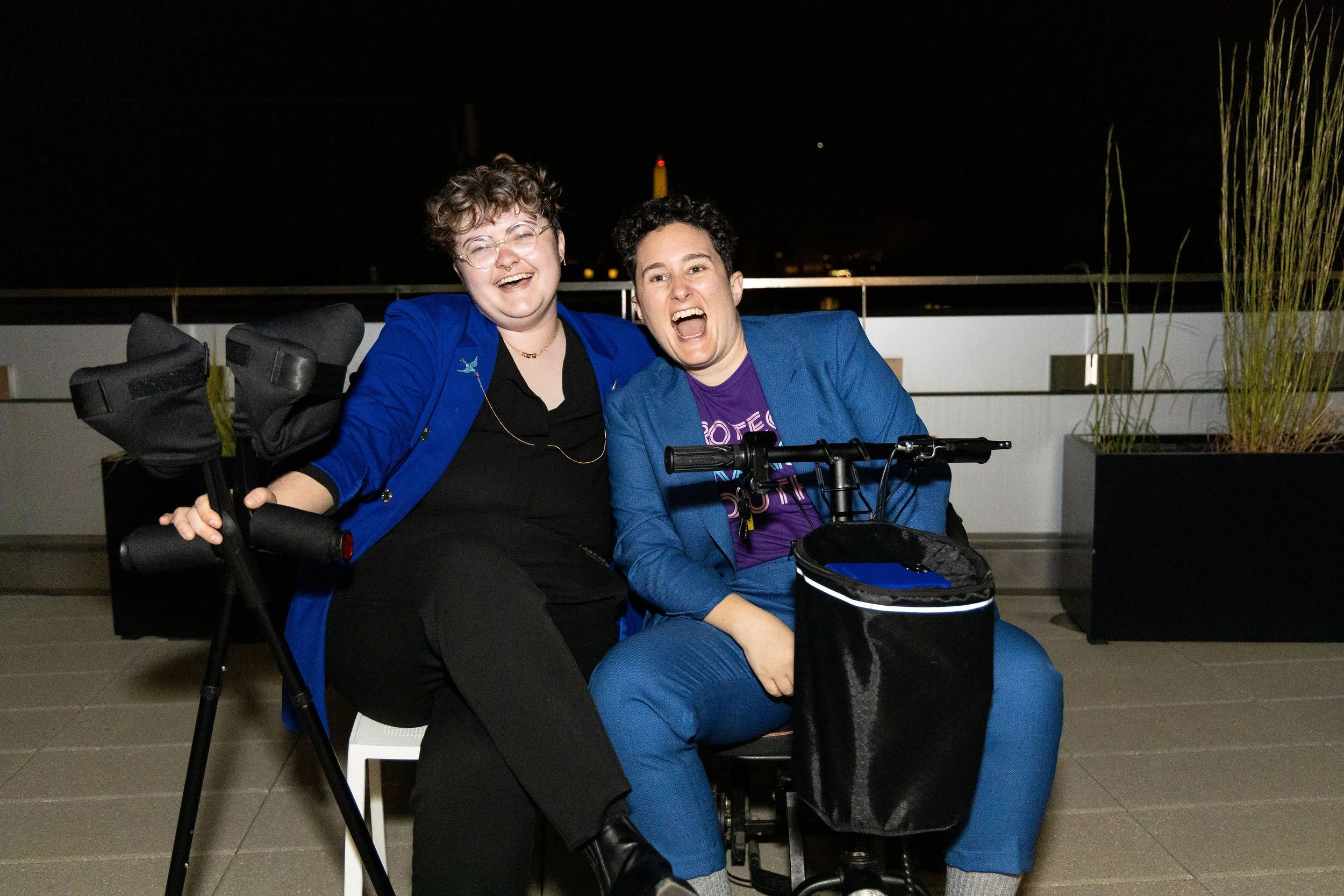 Two white people wearing black and blue formalwear sit on a rooftop and laugh together.