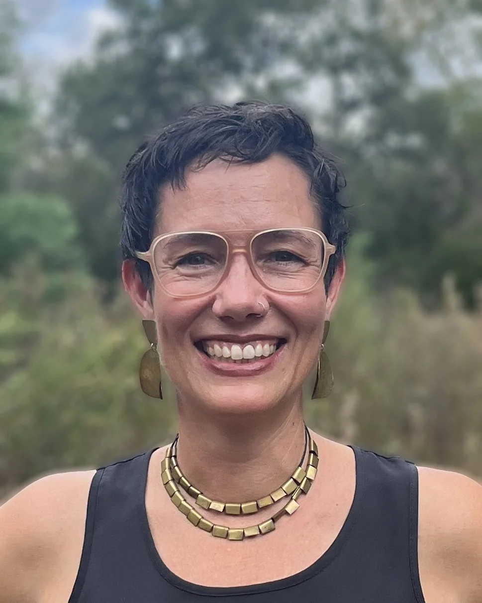 A smiling woman with short dark hair, wearing large glasses, gold earrings, a gold necklace, and a black tank top, outdoors in a natural setting with trees and a cloudy sky in the background.