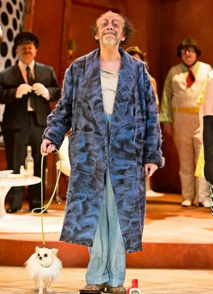 A man dressed as a disheveled old man holding a small dog on a leash, standing on a stage with people in costumes in the background.