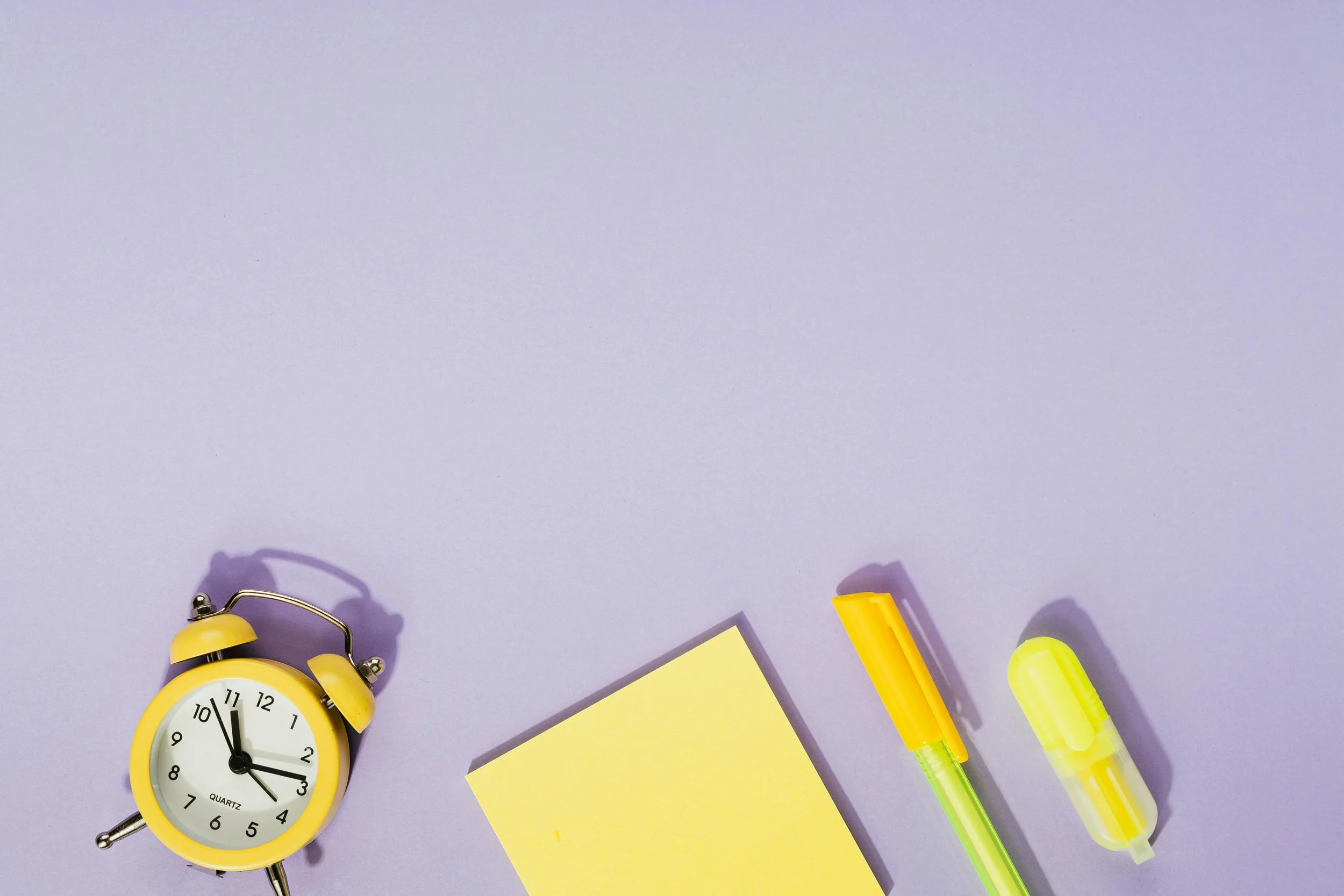 Yellow alarm clock, yellow notepad, yellow and green highlighter, yellow correction tape, on a lavender background represents opening timings of Legoland Billund resort.