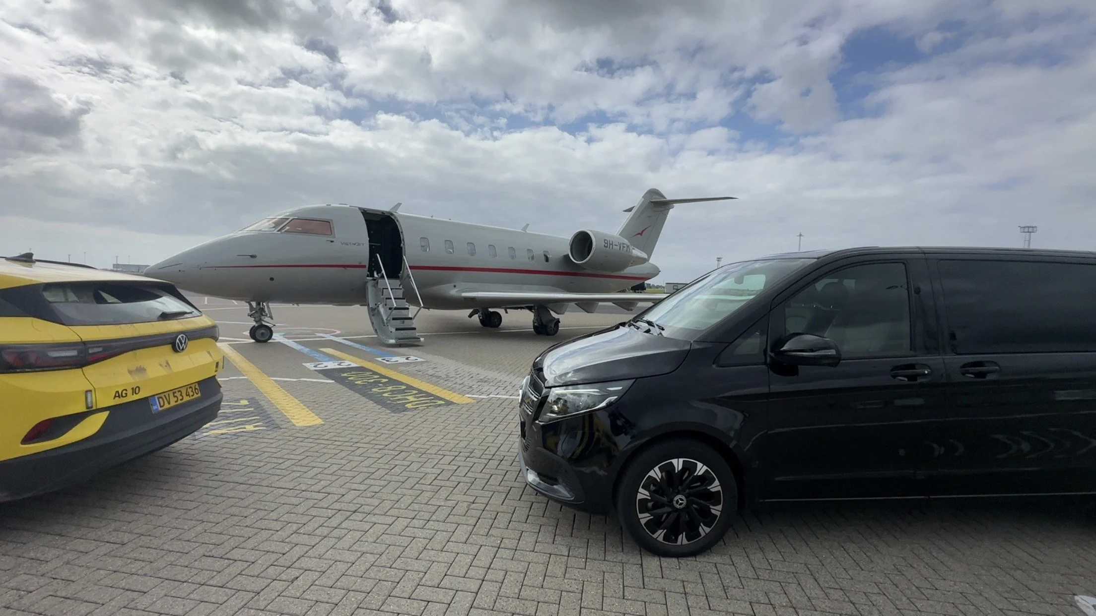 Private jet parked on the tarmac with open door, and a black Mercedes premium van nearby under a cloudy sky, ready to pick up a VIP guest for a private transfer.