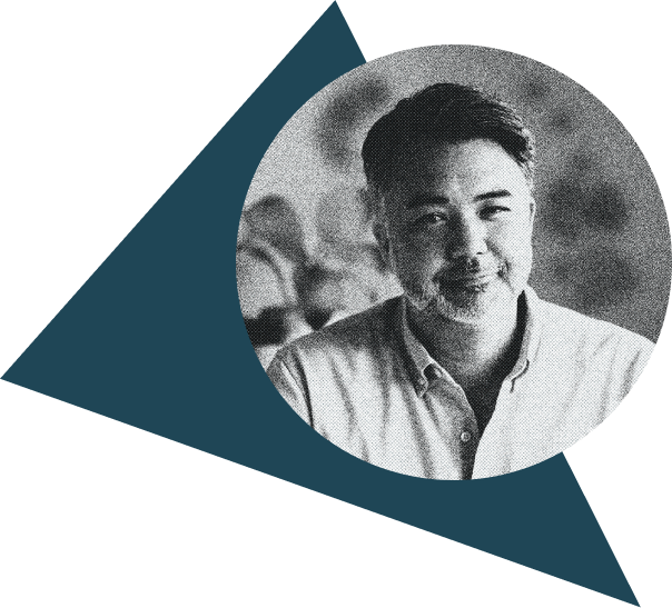 Black and white photo of a smiling man with dark hair and a beard, wearing a collared shirt, with blurred background and a geometric blue shape behind him.