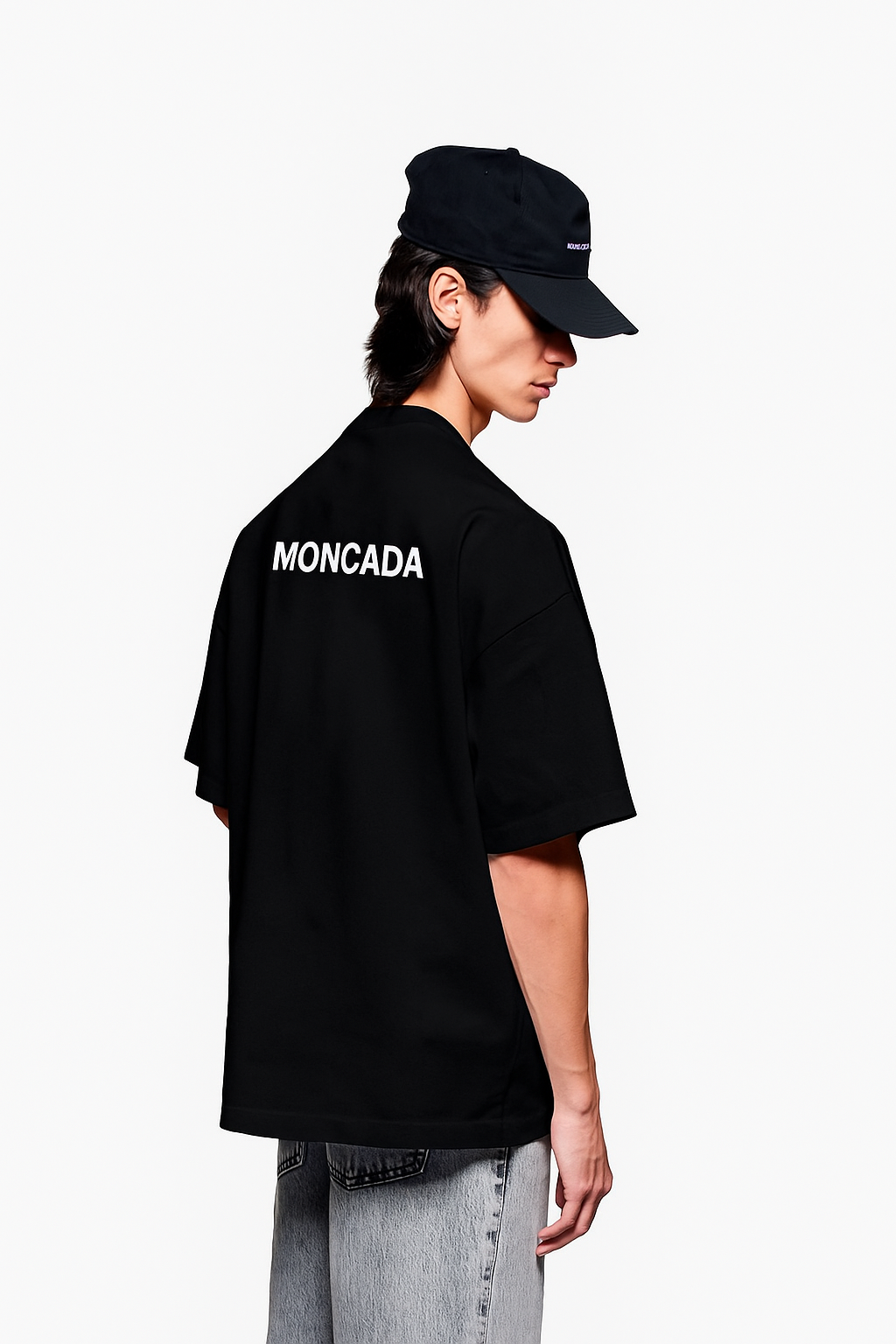 A young man with long dark hair wearing a black Moncada T-shirt and a navy blue cap, standing sideways against a plain white background.