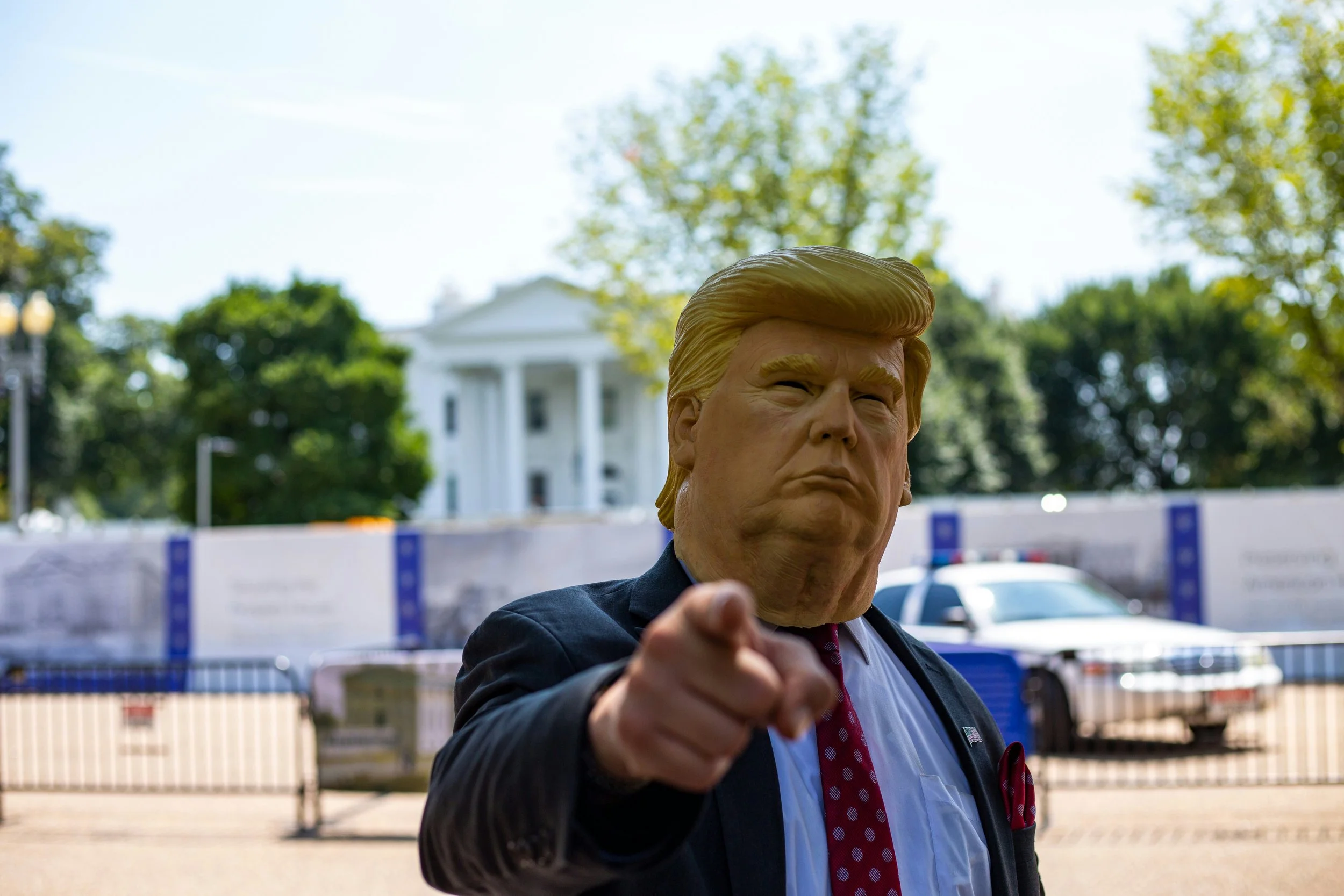 Person wearing a mask resembling Donald Trump, pointing at the camera, with the White House in the background.