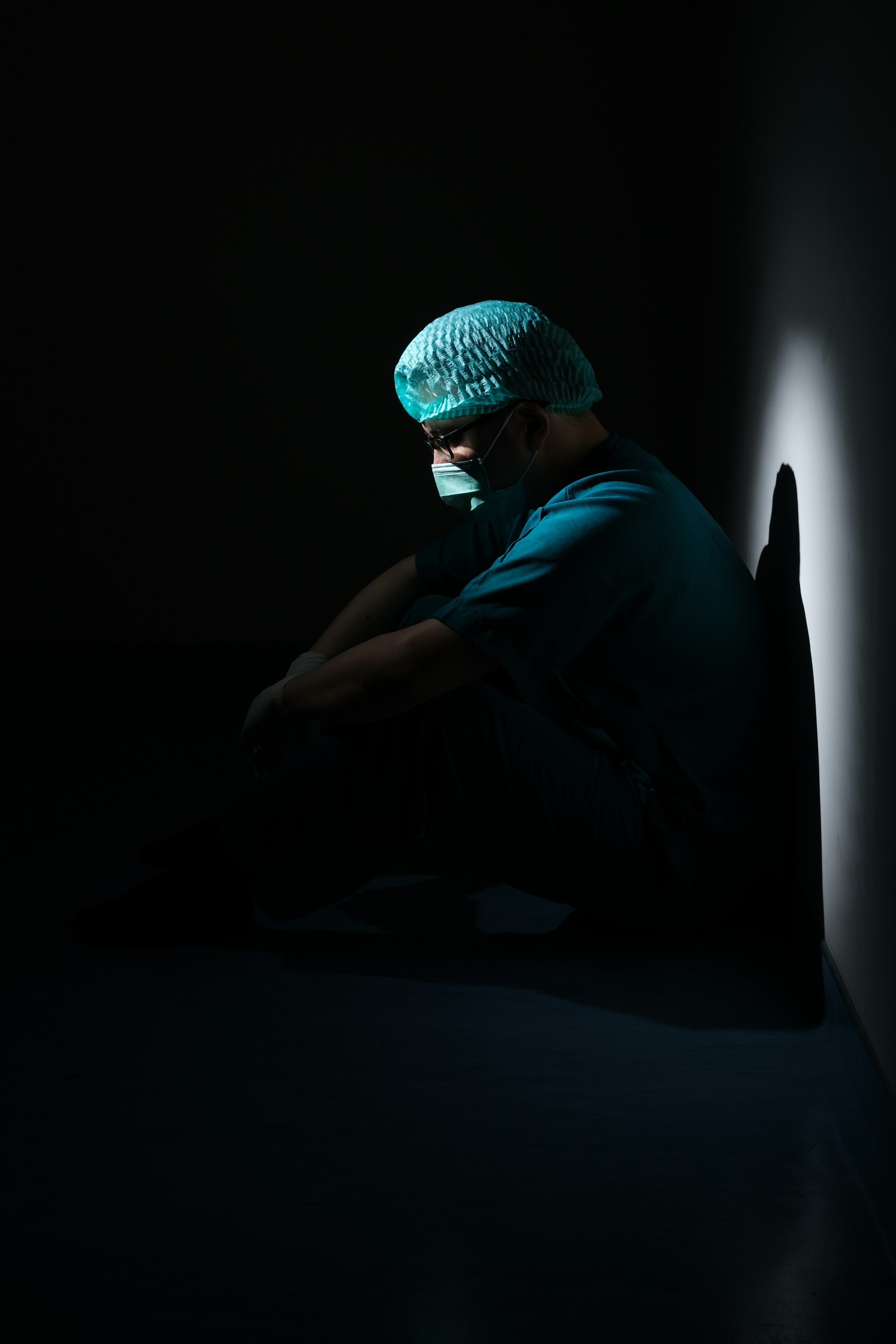 A doctor or nurse sitting on the floor in a dark room, wearing a surgical mask, cap, and glasses, with their head bowed and an exhausted posture, illuminated by a single light.