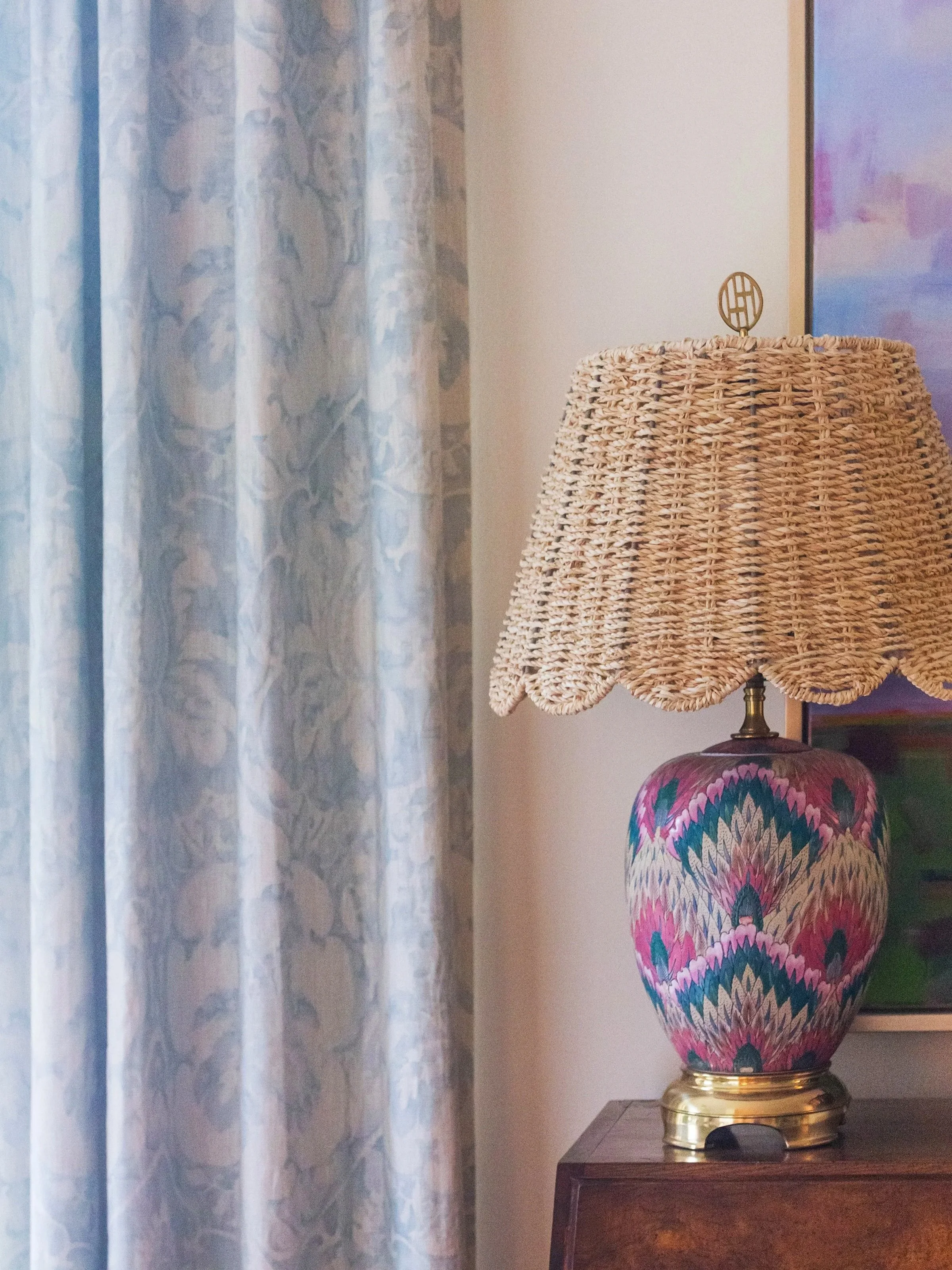 Decorative table lamp with a woven wicker shade, a colorful patterned vase with a floral design, positioned on a wooden table near a curtain and wall art.