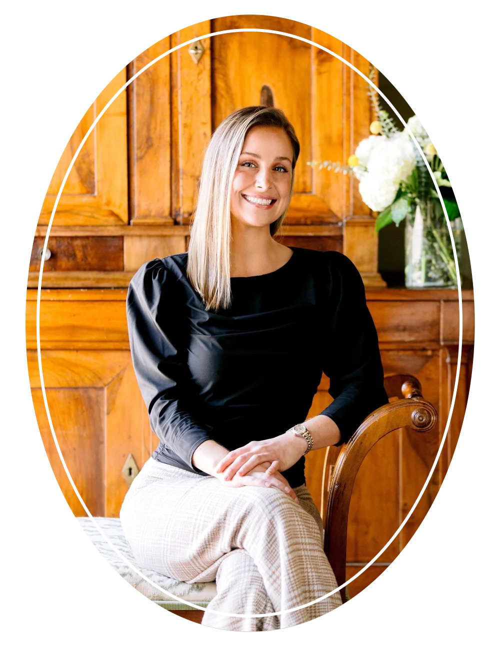Smiling woman with long blonde hair, wearing a black top and beige checkered pants, seated in a wooden chair with a wooden background and a vase of white flowers in the background.