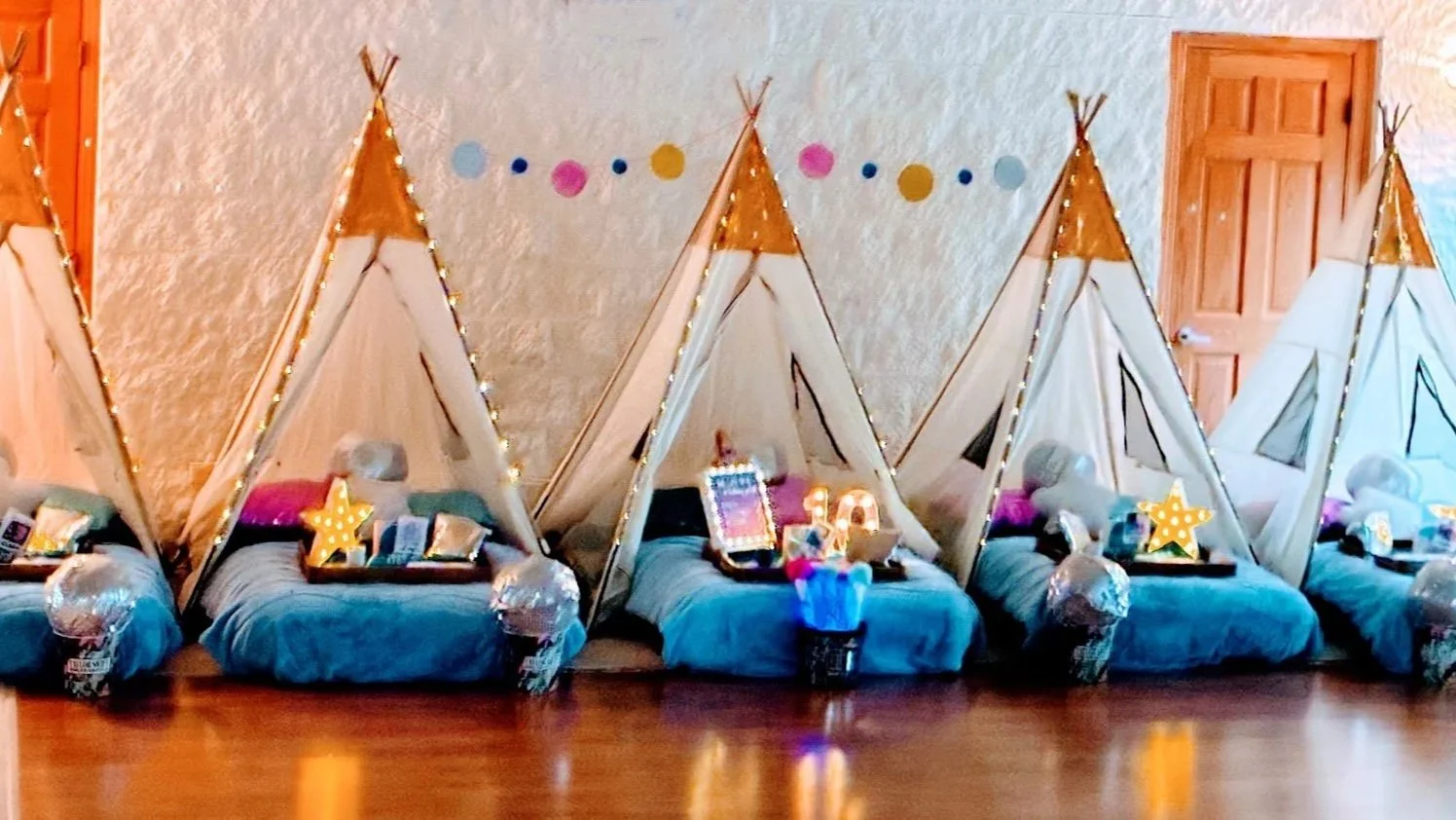 Indoor scene with five teepee tents decorated for a children's sleepover or party, each with fairy lights, plush pillows, stuffed animals, star-shaped lamps, and trays of snacks and drinks, against a beige textured wall with colorful dot garland.
