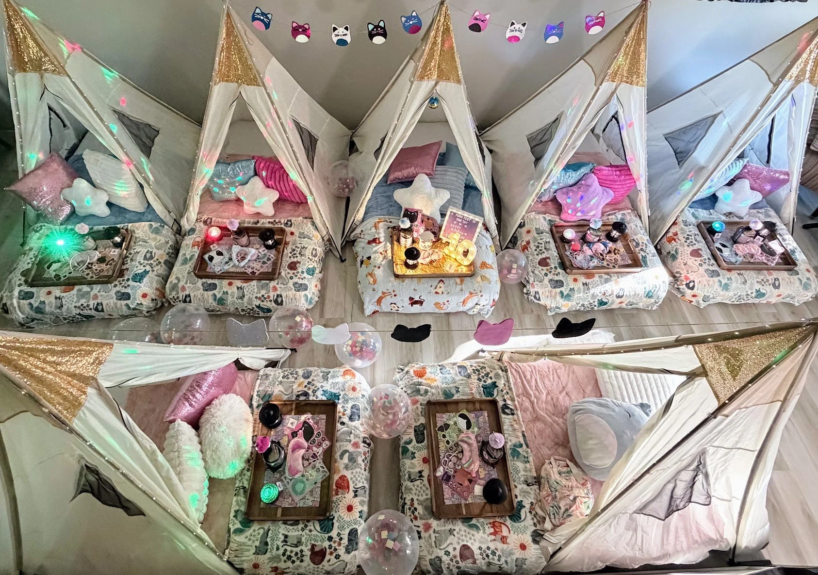 Top-down view of a cozy indoor space with eight small beds, each with a canopy, colorful pillows, and decorations. There are decorative panels with cat face designs hanging from the ceiling, and birthday or celebration items such as balloons, candles, and small treats are placed on trays and surrounding the beds.