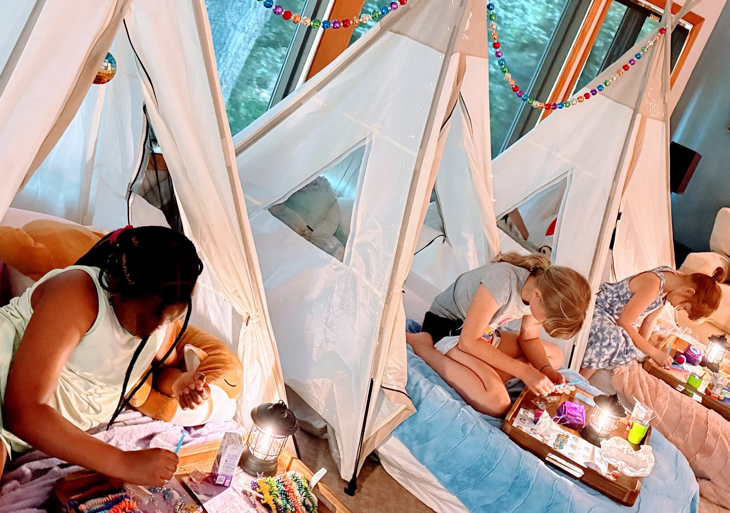 Three girls sitting in tents indoors, engaged in arts and crafts by candlelight, with colorful rainbow birthday decorations hanging above them.