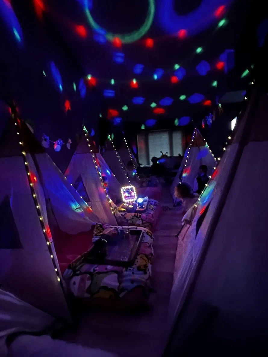 A dark room with colorful laser light projections on the ceiling, and small tents with string lights arranged inside. There are children sitting near the tents, and a tray and a few items on the floor.
