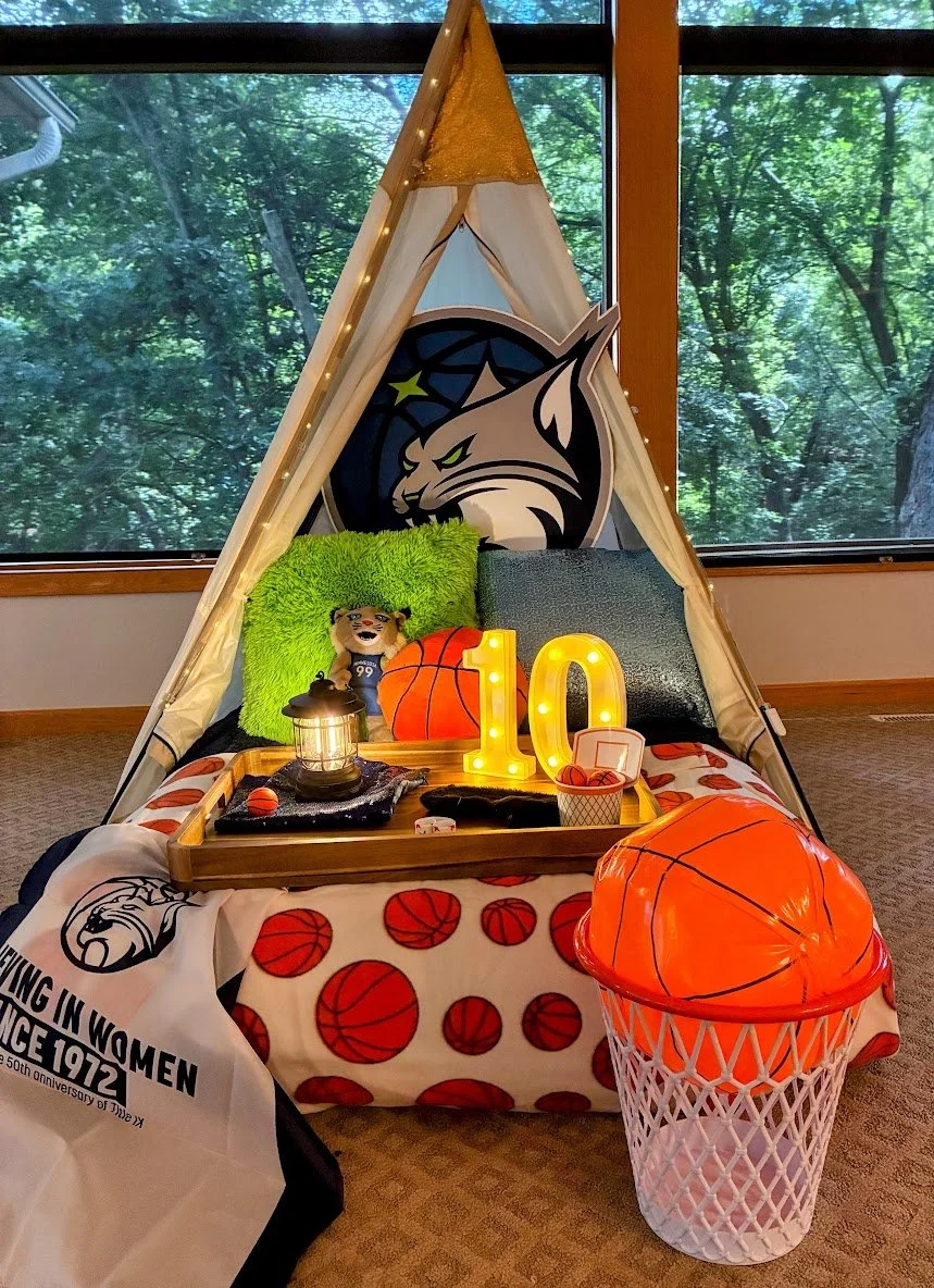 Indoor teepee decorated for a basketball Lynx birthday with basketball theme, illuminated number 10, a basketball toy, plush pillow, lantern, and a hoop shaped basket with inflatable basketball.