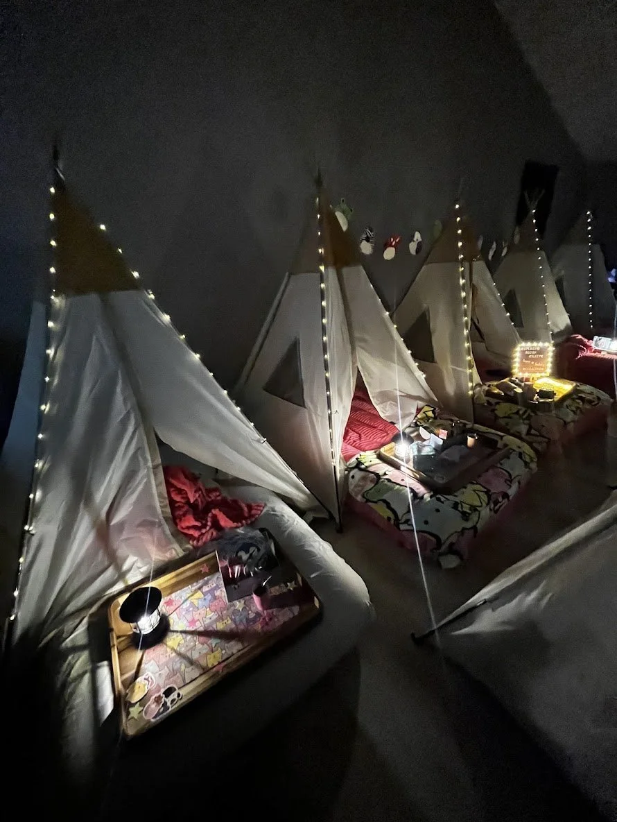 Five beds with white fabric canopies, each decorated with string lights, in a dimly lit room. Each bed has a tray with snacks and drinks, and the beds are positioned side by side.
