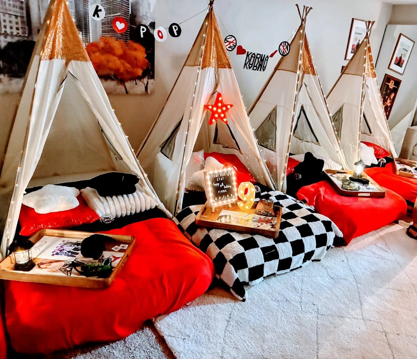 Kids party set up: a small kids tent decorated with a red star-shaped light hanging from the top, a plush star-shaped pillow, and sparkly pillows.  A tray shows K-Pop star Lee Know from Stray Kids. A black and white checked blanket is on bed.