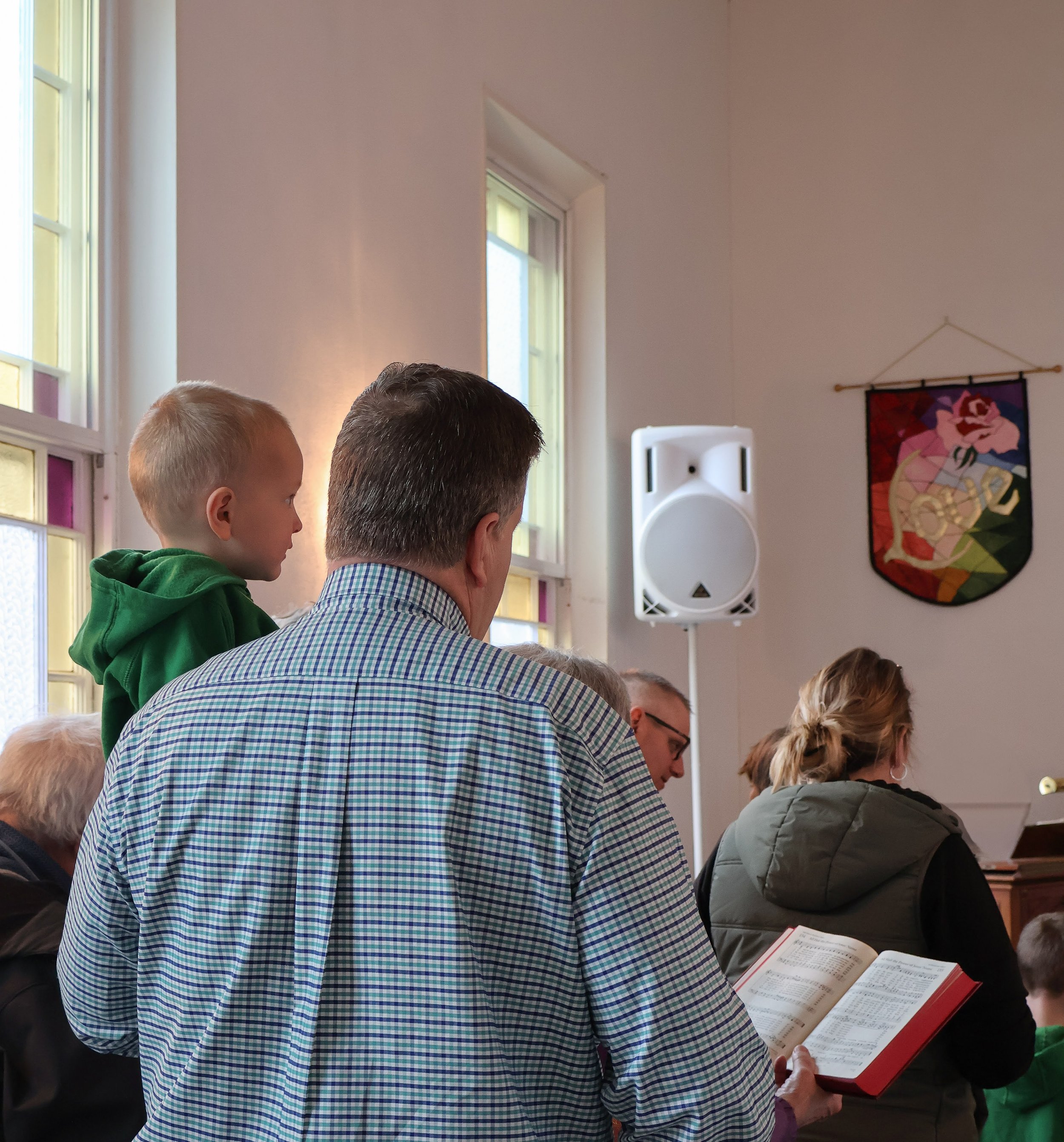People gathered in a room with stained glass windows, some reading from hymnals or prayer books, and a shield with a lion emblem on the wall.