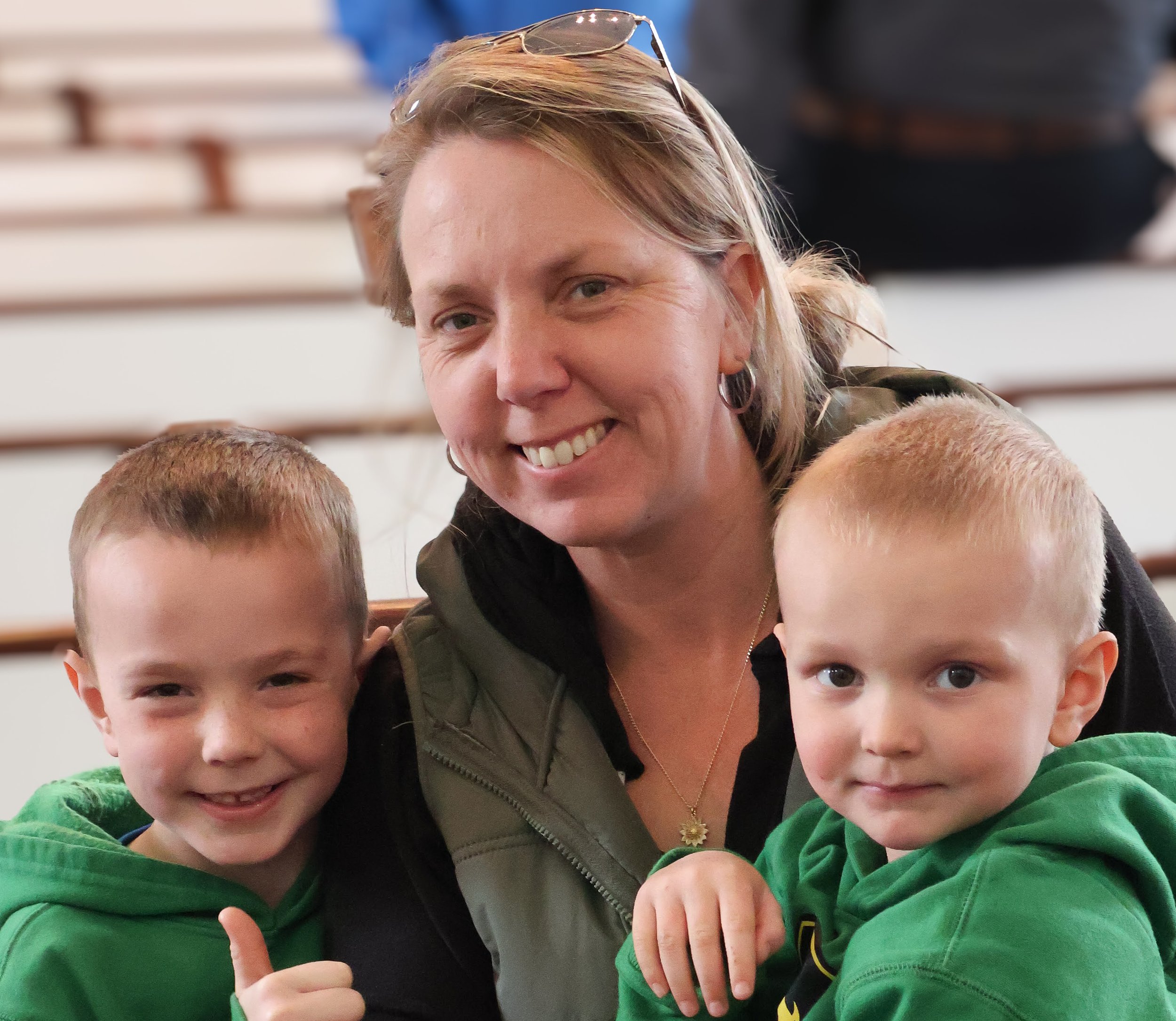A woman smiling with two young boys, all wearing green hoodies, sitting in a gymnasium with bleachers in the background.