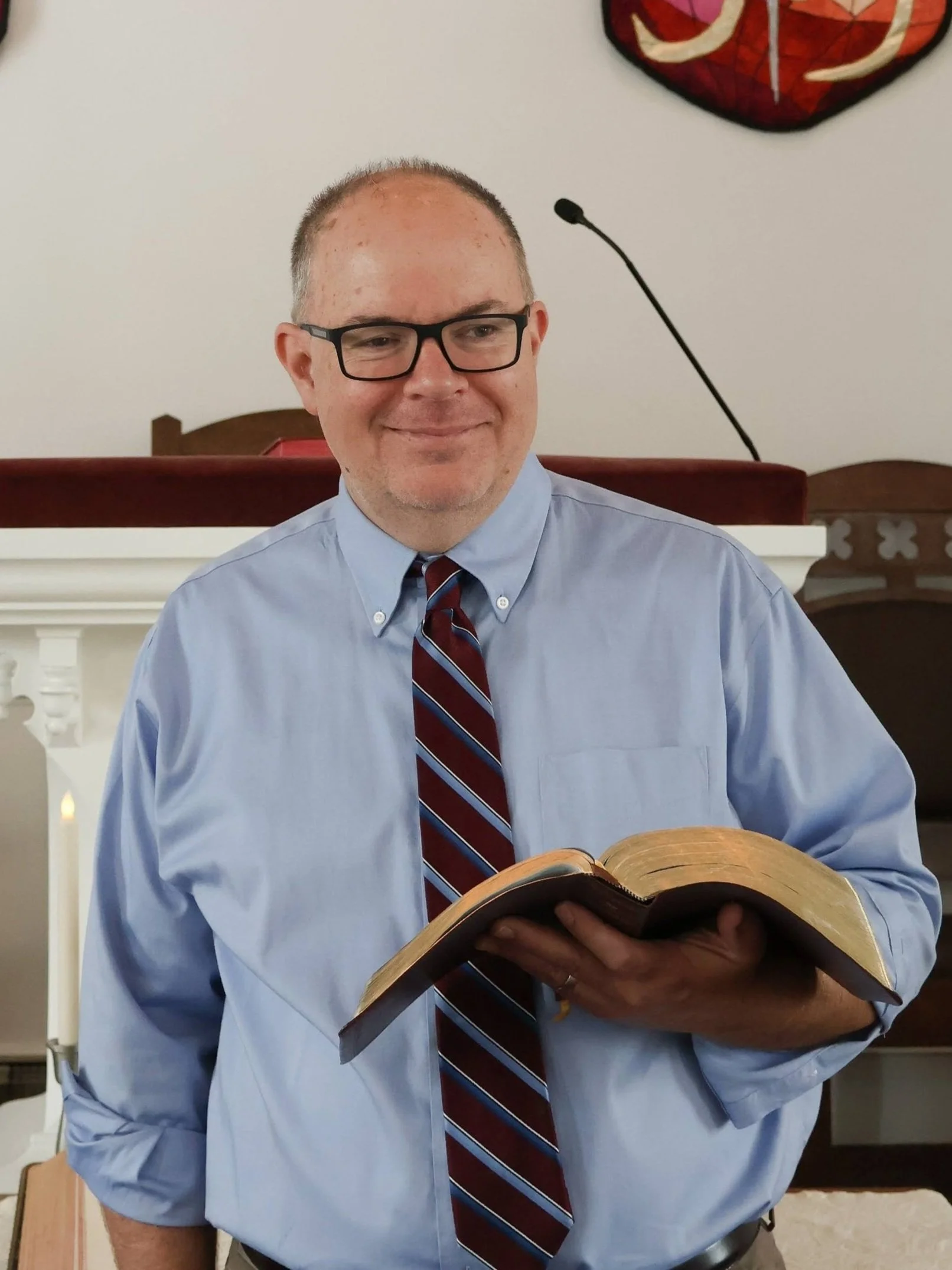A man with glasses, a light blue shirt, and a striped red and blue tie holding an open Bible inside a church.