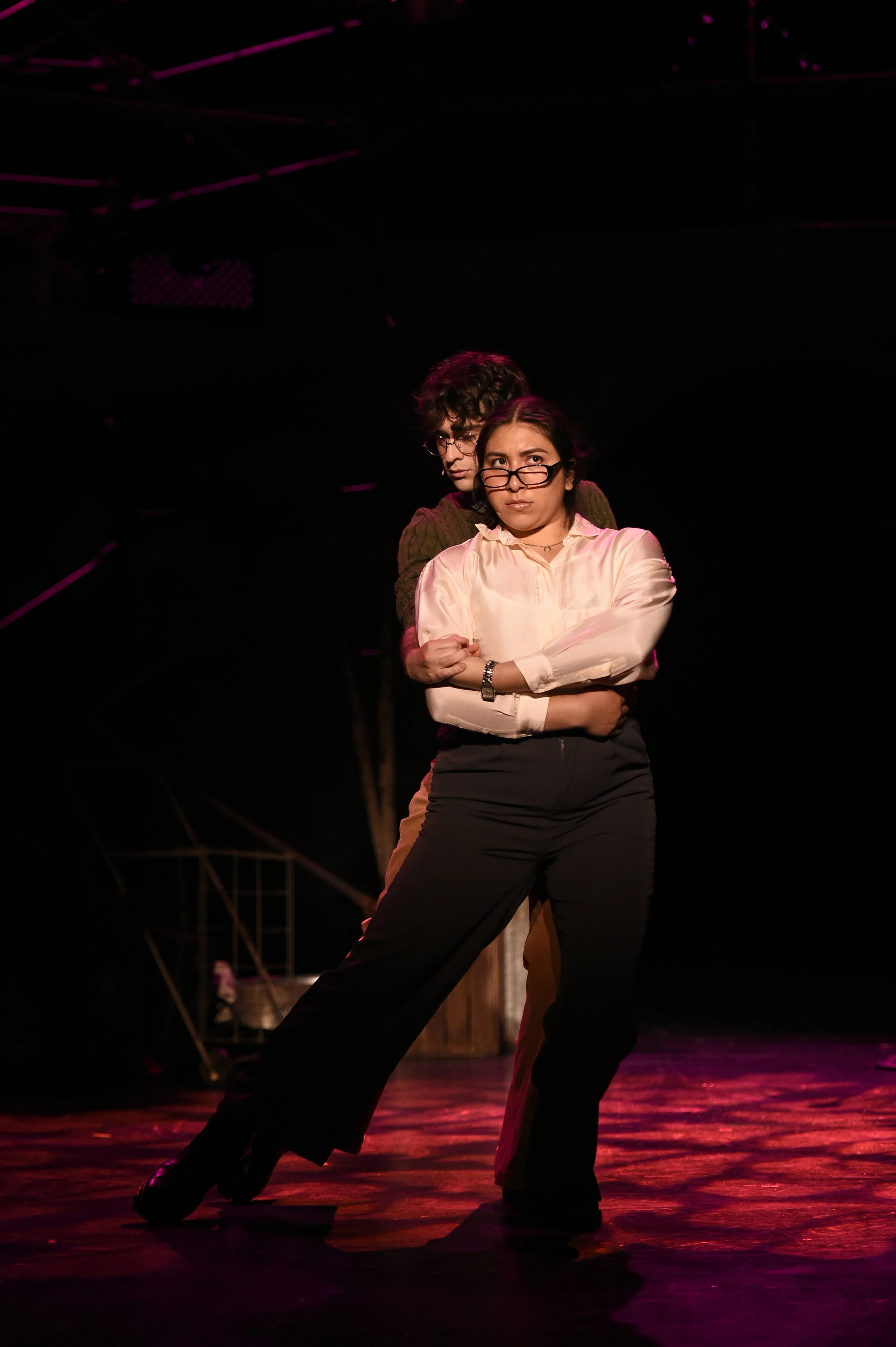Two actors performing on stage, one woman in a white blouse and black pants with glasses, the other man behind her, softly embracing her.