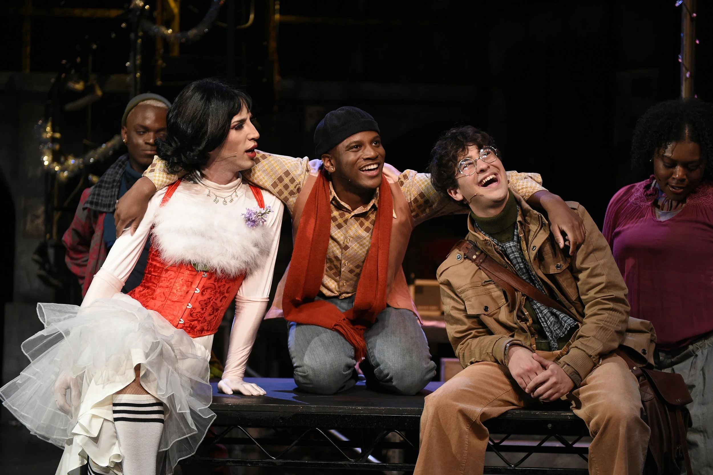 Group of actors on stage during a theatrical performance, dressed in colorful costumes, with a black background and stage props.