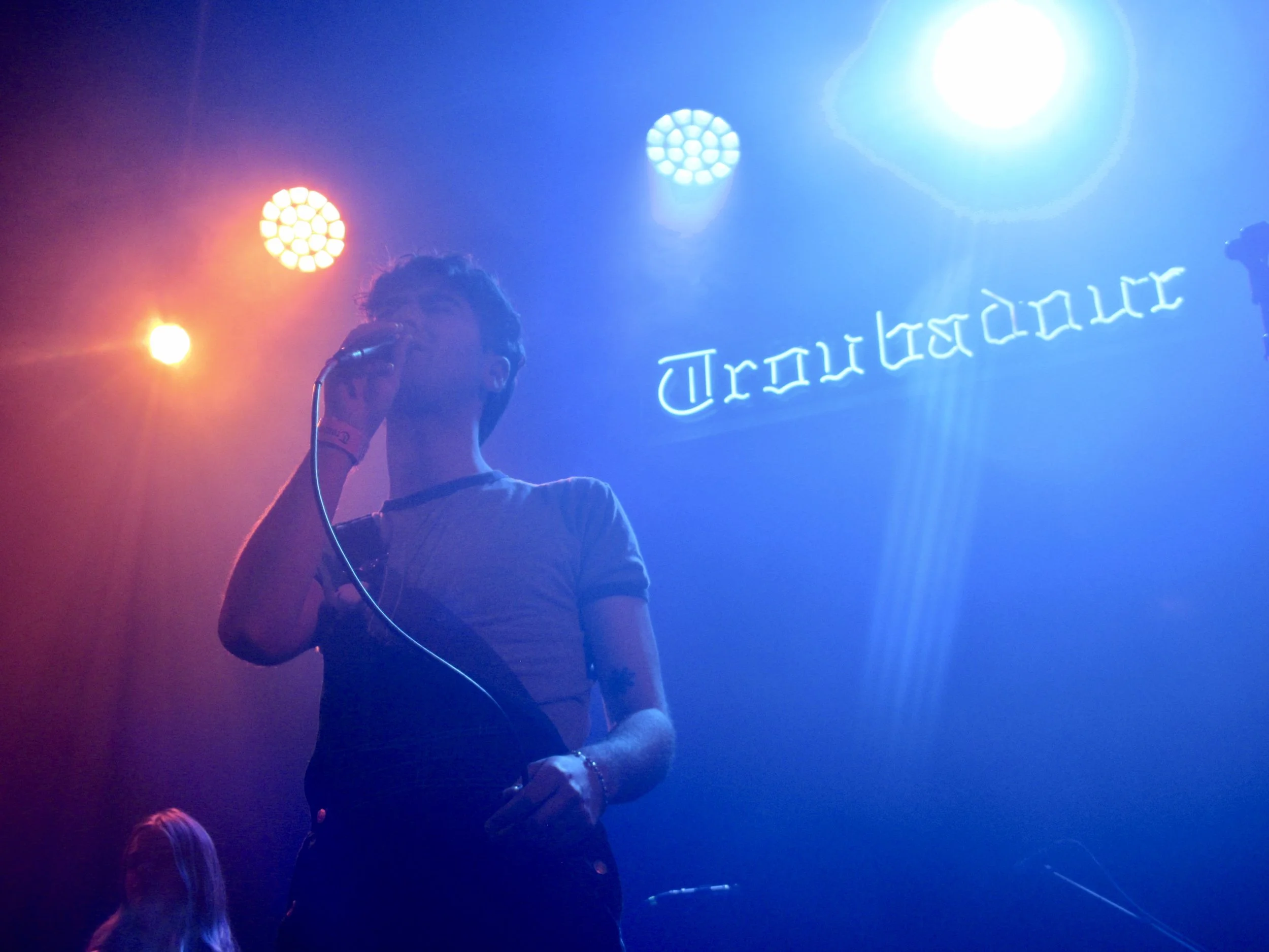 A person singing into a microphone on stage with multicolored stage lights and a bright sign that reads 'Troubadour' in the background.