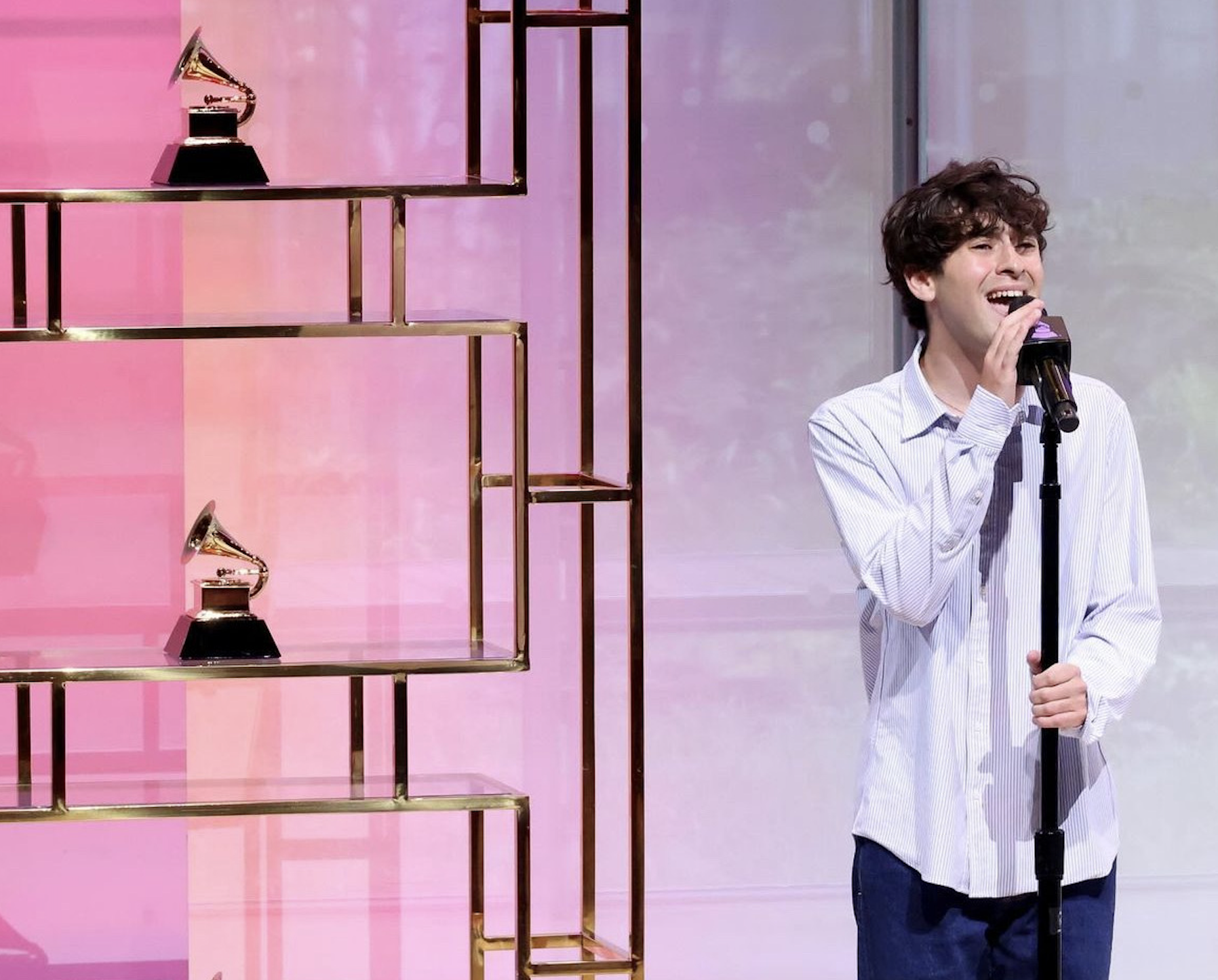 A young man with curly brown hair singing into a microphone on stage, standing in front of a pink background with a gold shelving unit holding two gramophone trophies.