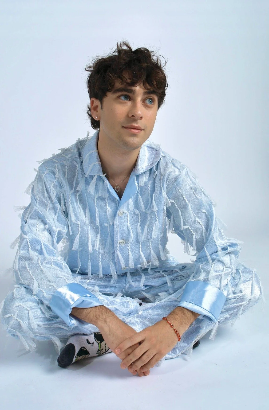 A young man with curly brown hair and blue eyes sitting on the floor, wearing a light blue, frayed, patterned shirt and matching pants, with a red bracelet on his wrist and patterned socks.