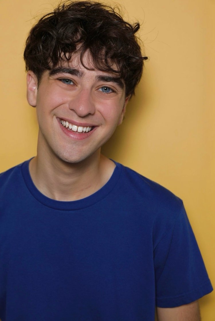 A young man with curly dark hair smiling and wearing a blue T-shirt, standing against a yellow background.