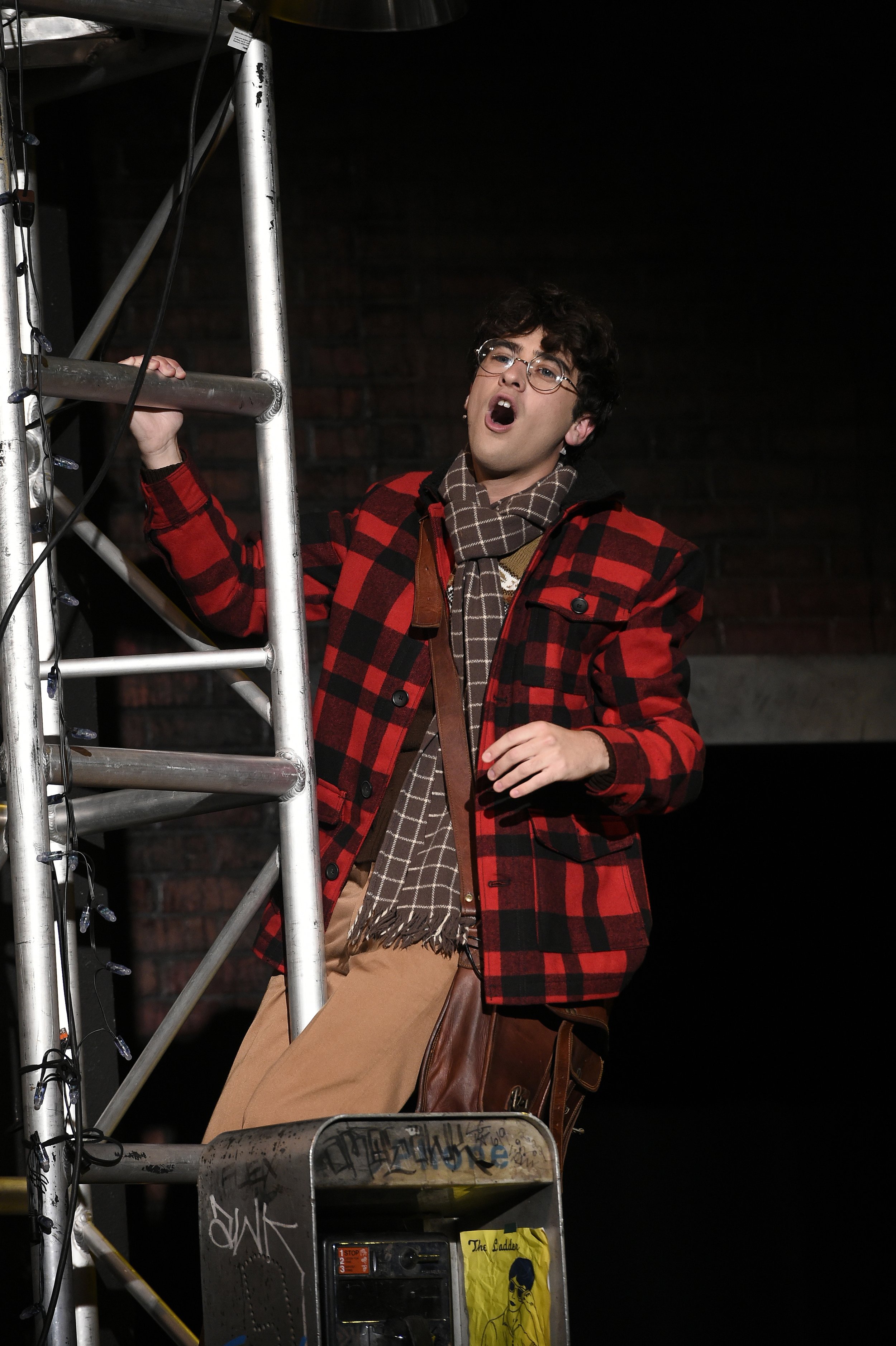 A young man wearing glasses, a red and black plaid jacket, and a brown scarf sings on stage near a metal scaffold, with a dark brick background.
