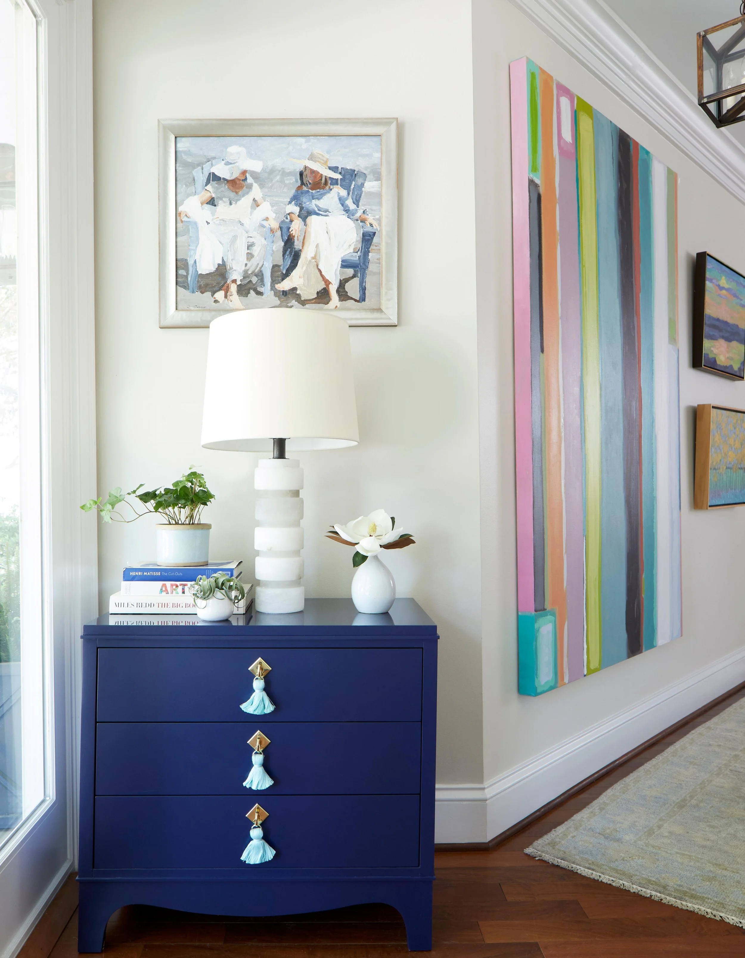 Interior of a living space featuring a dark blue dresser with three drawers, decorated with tassel pulls, topped with a white table lamp, a small potted plant, a stack of books, a white vase with a flower, and artwork on the wall, including a colorfu