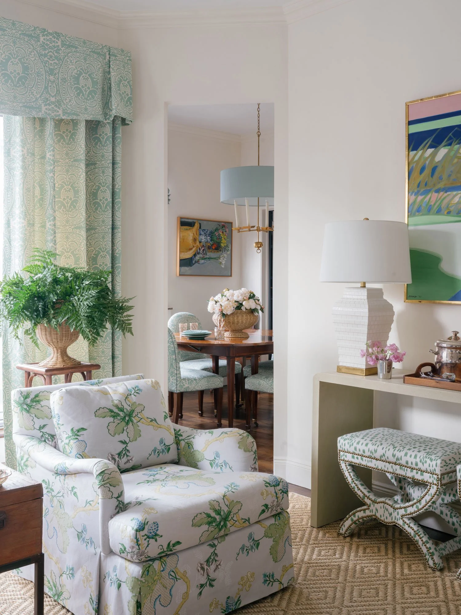 Living room view of New York City apartment by Elizabeth Hartz Interior Design showing floral upholstered accent chair, patterned aqua drapery, textured jute rug, and console with bar styling, opening into dining area with custom upholstered dining c