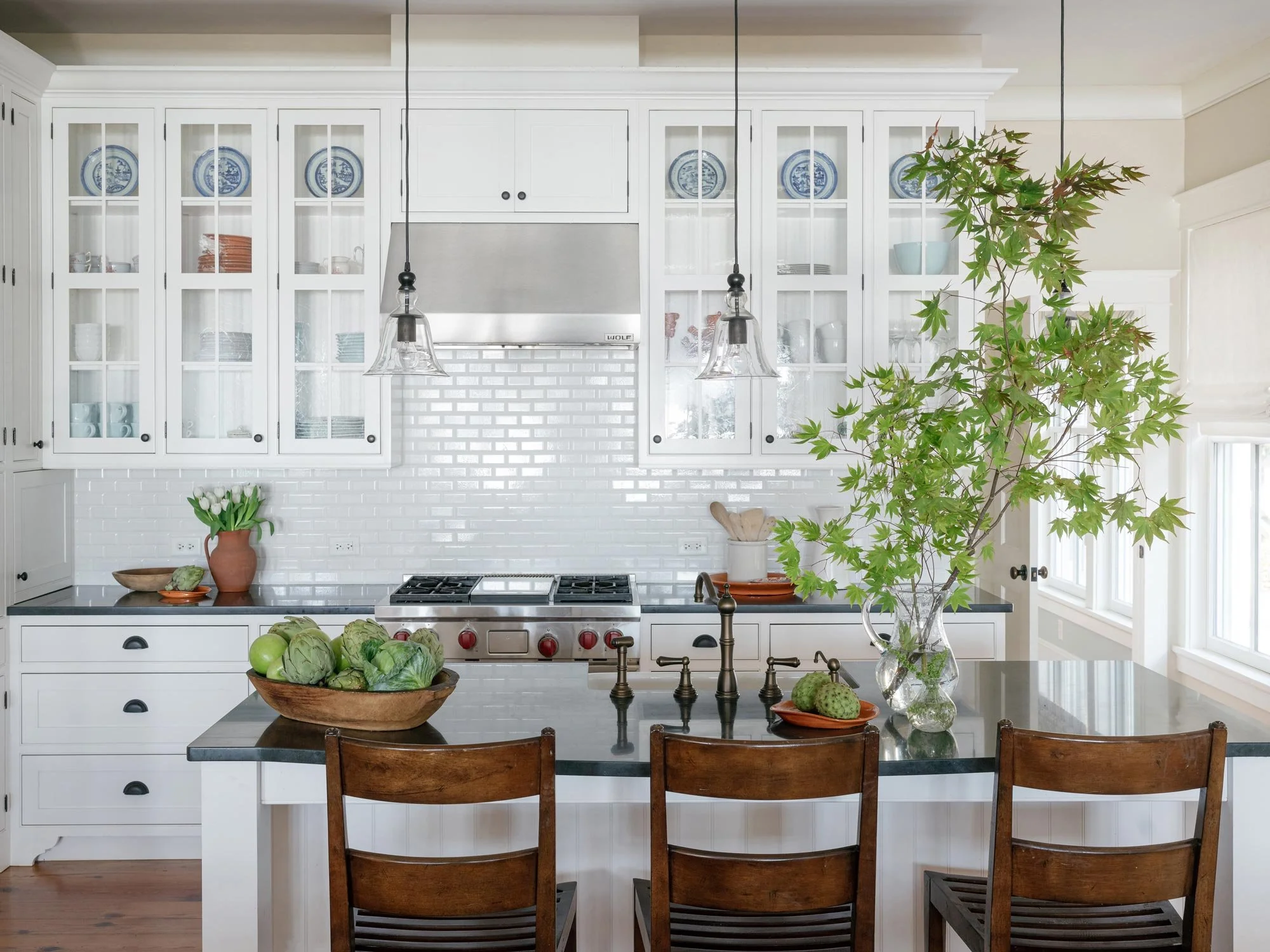 Light-filled kitchen designed by Elizabeth Hartz Interior Design at The River home in Kilmarnock, Virginia. Features white cabinetry with glass doors, subway tile backsplash, and a dark stone island countertop with wooden stools and fresh greenery fo