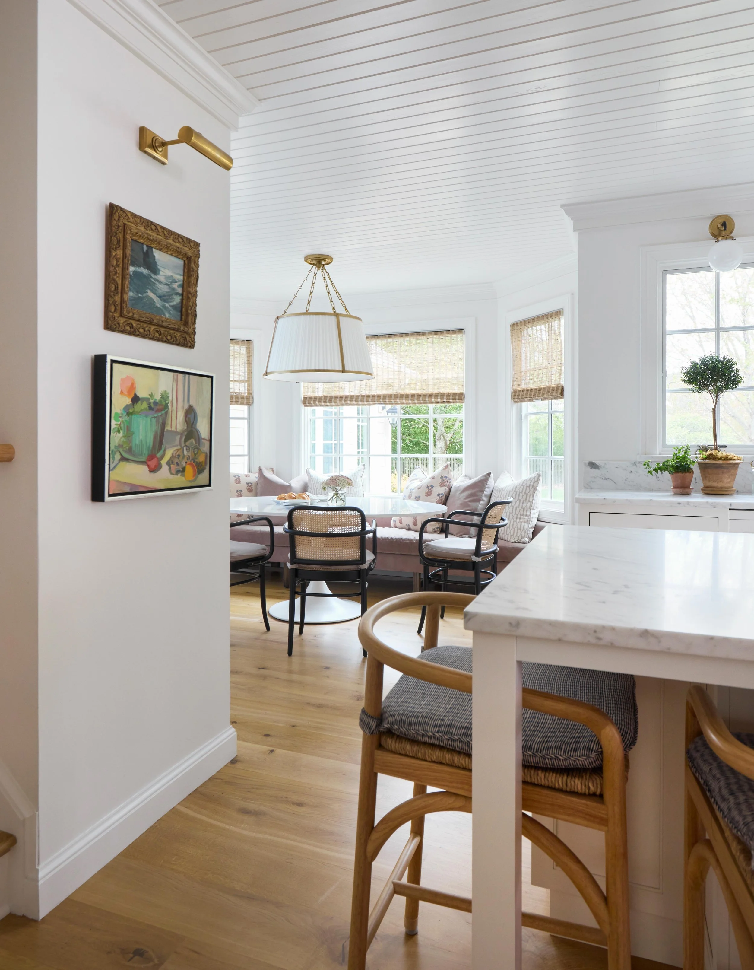 Bright dining and living area with white walls, wooden floors, large windows with woven blinds, a round table surrounded by chairs, and a cozy corner with cushions and plants.